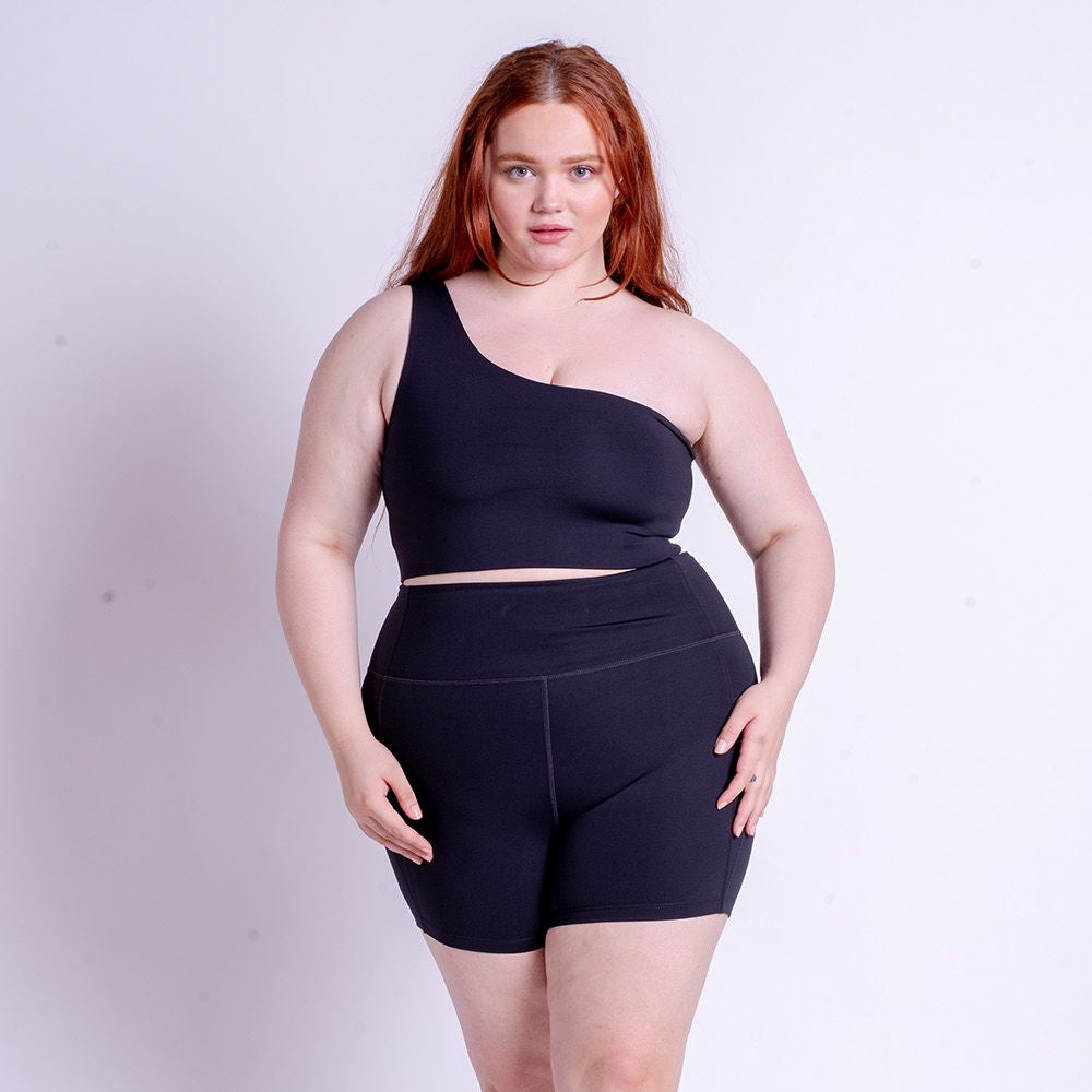 Frau mit roten Haaren trägt ein schwarzes, asymmetrisches Top und passende Shorts vor einem weißen Hintergrund.