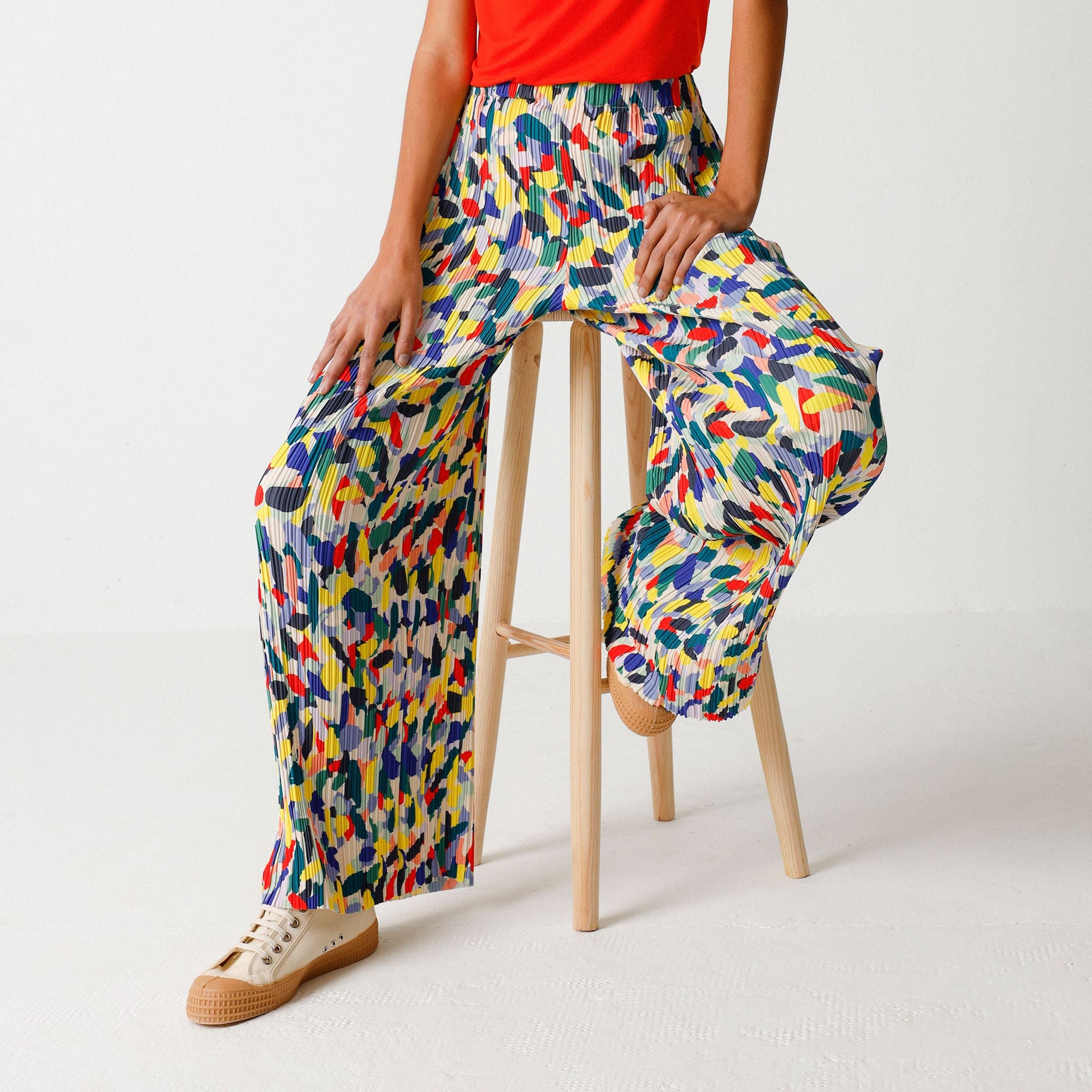 Eine Person sitzt auf einem Hocker und trägt eine bunte, gemusterte Hose und ein rotes Oberteil.