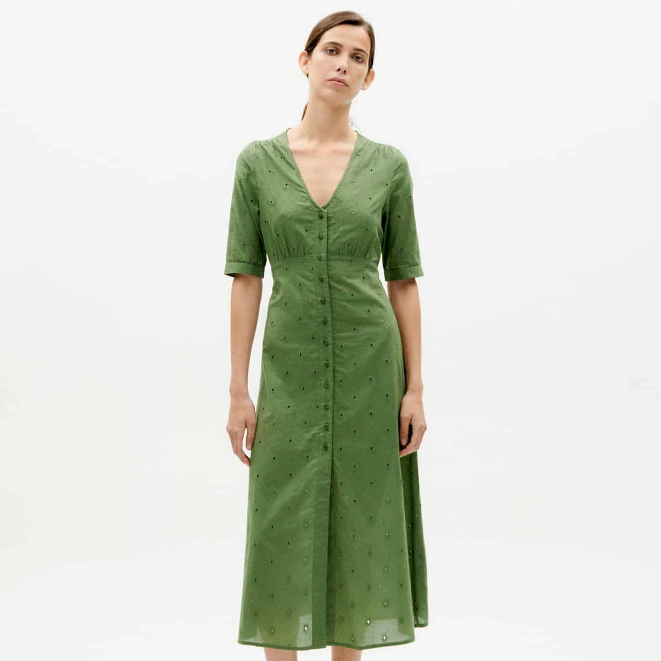 Frau in grünem, knielangem Kleid mit kurzen Ärmeln und Knöpfen, steht vor einem weißen Hintergrund.