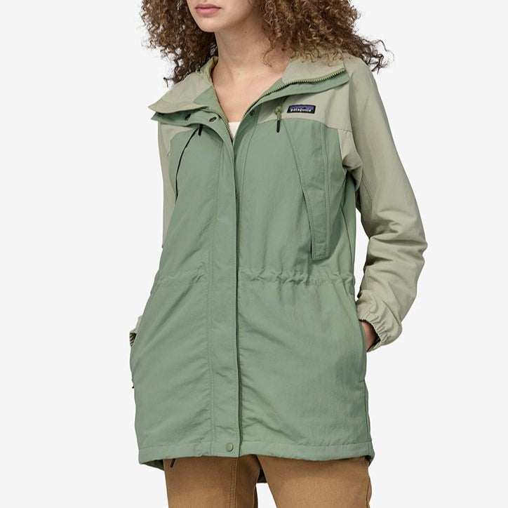 Frau trägt eine zweifarbige grüne Jacke mit Kapuze und hat die Hände in den Taschen.