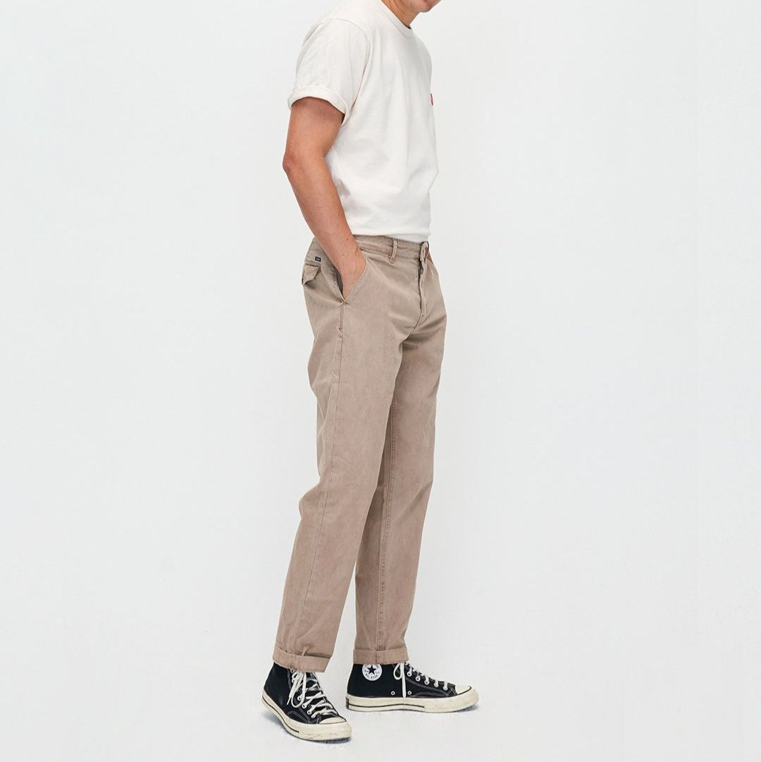 Ein Mann in beiger Hose und weißen T-Shirt steht seitlich und trägt schwarze hochgeschnürte Sneaker.