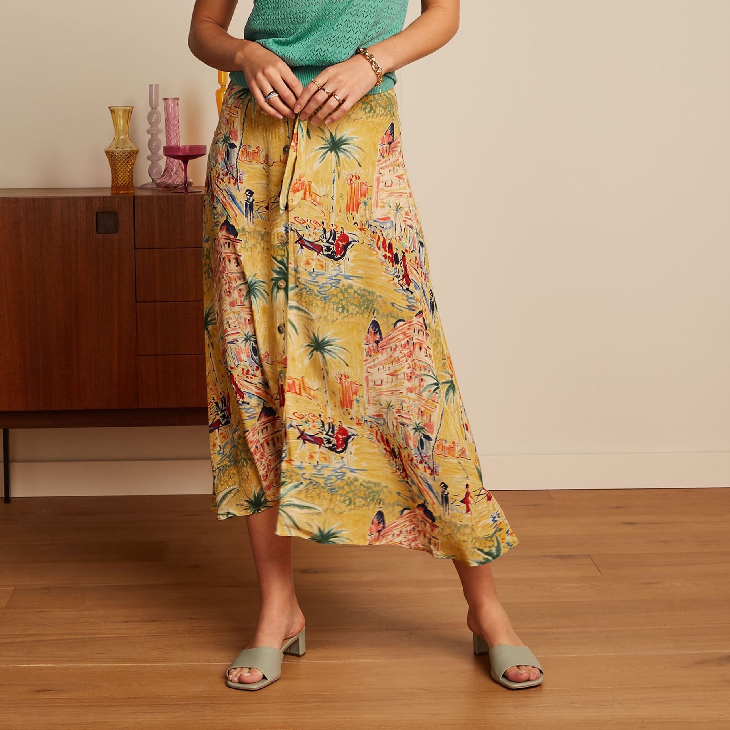 Frau in grünem Oberteil und buntem Rock mit Stadtmotiv steht neben einem Sideboard auf Holzboden.