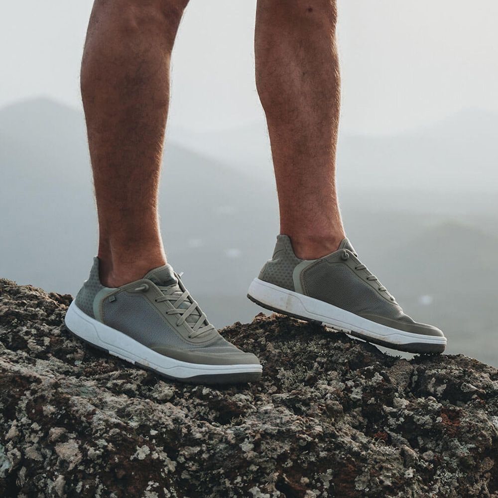 Nahaufnahme von Beinen in grauen Sneakers, die auf einem Felsen stehen, mit verschwommenem Hintergrund von Bergen.