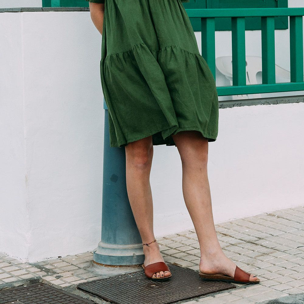 Person in a green dress and brown sandals leaning against a blue pole on a tiled sidewalk.