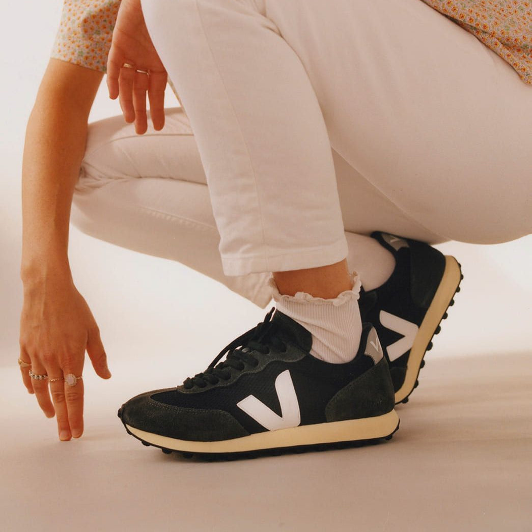 Person in weißen Hosen hockt und trägt schwarze Sneaker mit weißem Logo.