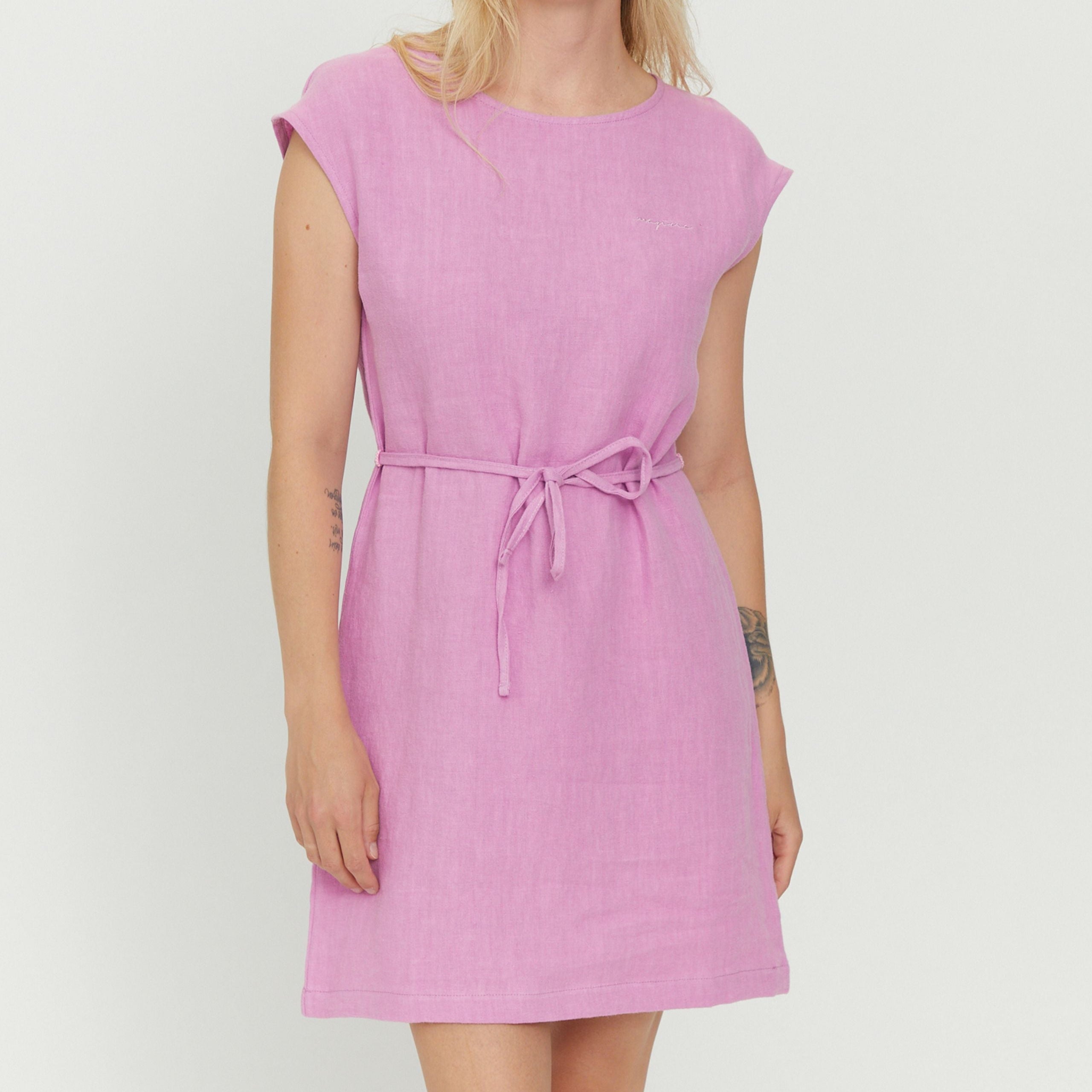 Frau trägt ein lässiges, lila Kleid mit Gürtel in der Taille, ihre Arme sind sichtbar, und sie hat ein Tattoo auf dem Arm.