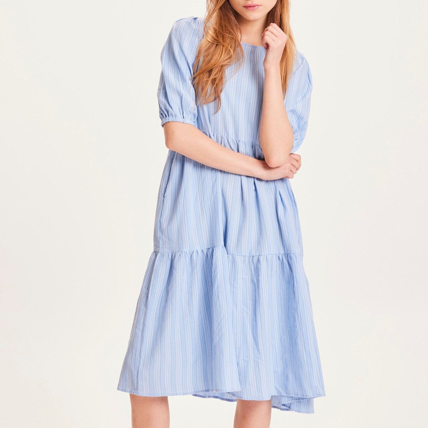 Eine Person trägt ein hellblaues, gestreiftes Kleid mit kurzen Ärmeln vor neutralem Hintergrund.