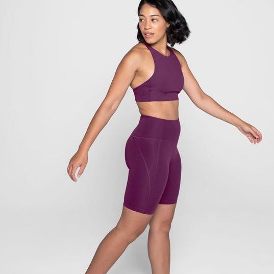 Frau in lila Sportbekleidung mit Top und Shorts vor grauem Hintergrund.