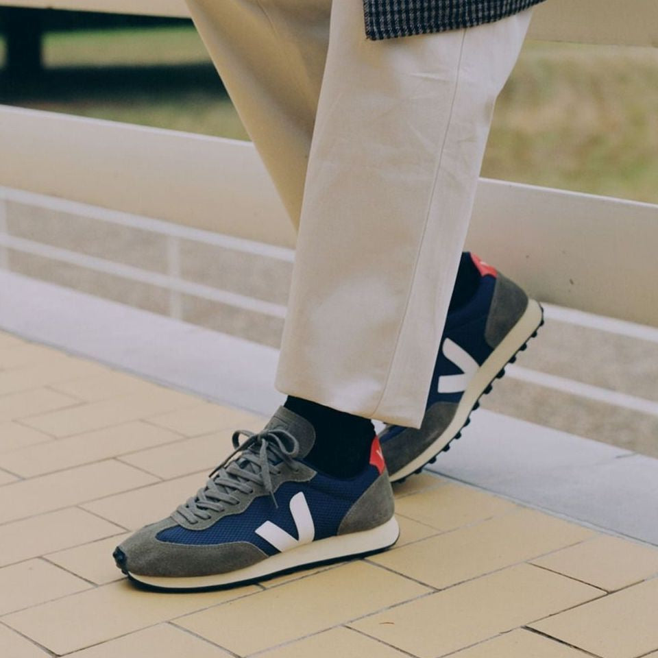 Person in weißen Hosen und schwarzen Socken trägt graue und blaue Sneakers mit weißem Logo, auf gelbem Fliesenboden.