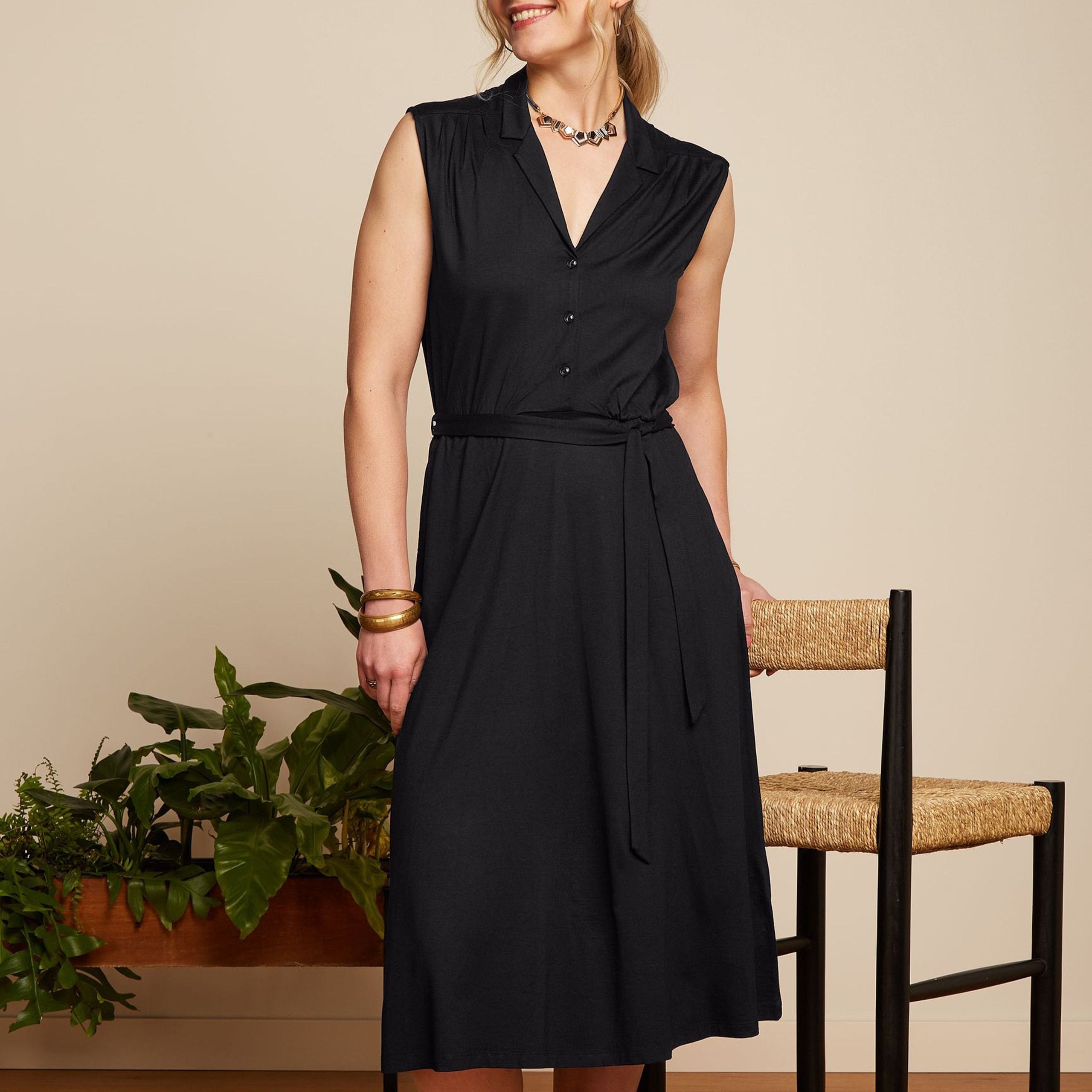 Frau in schwarzem ärmellosen Kleid steht neben einem Stuhl, mit Pflanzen im Hintergrund.