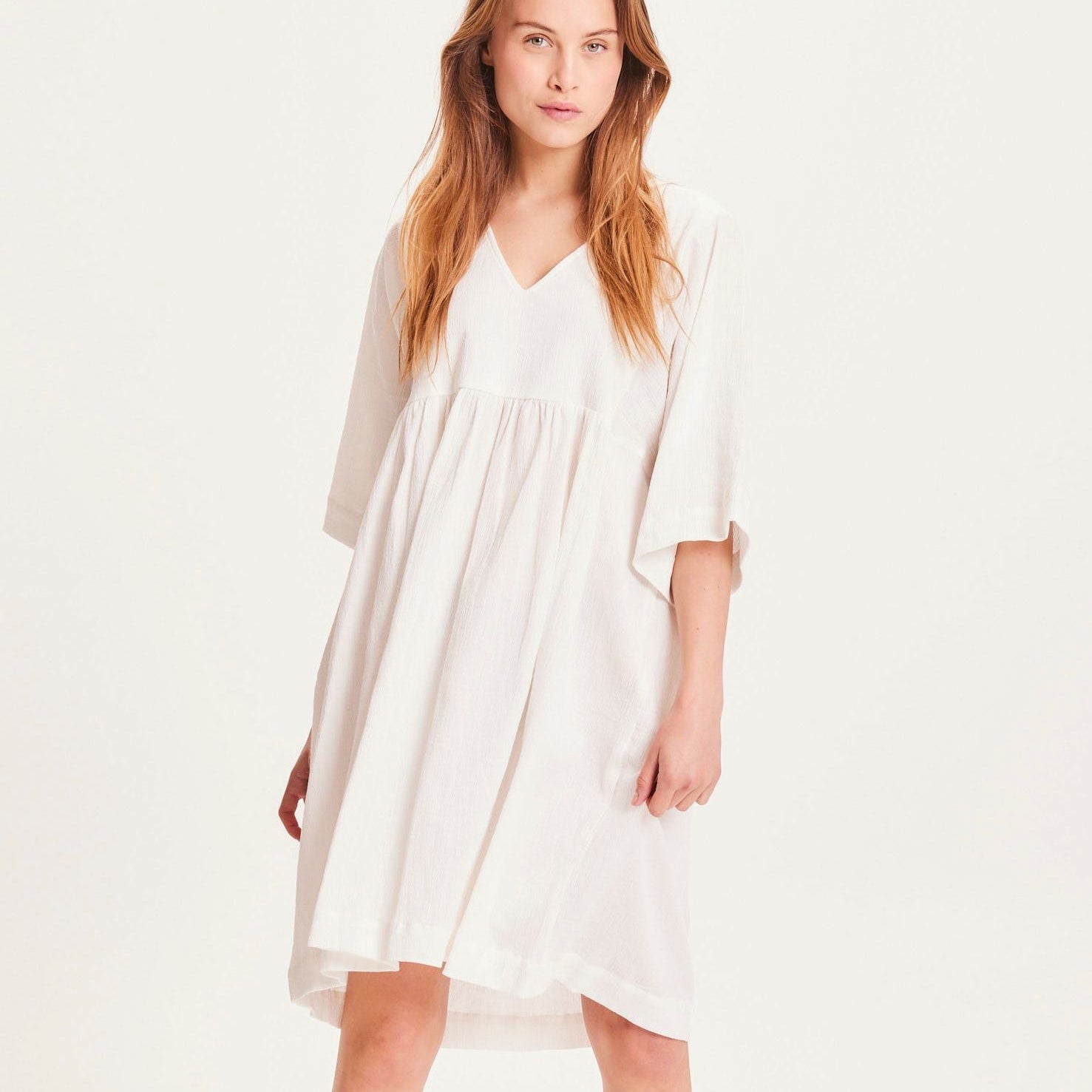 Frau in einem weißen, fließenden Kleid mit V-Ausschnitt und halblangen Ärmeln, stehend vor einem hellen Hintergrund.