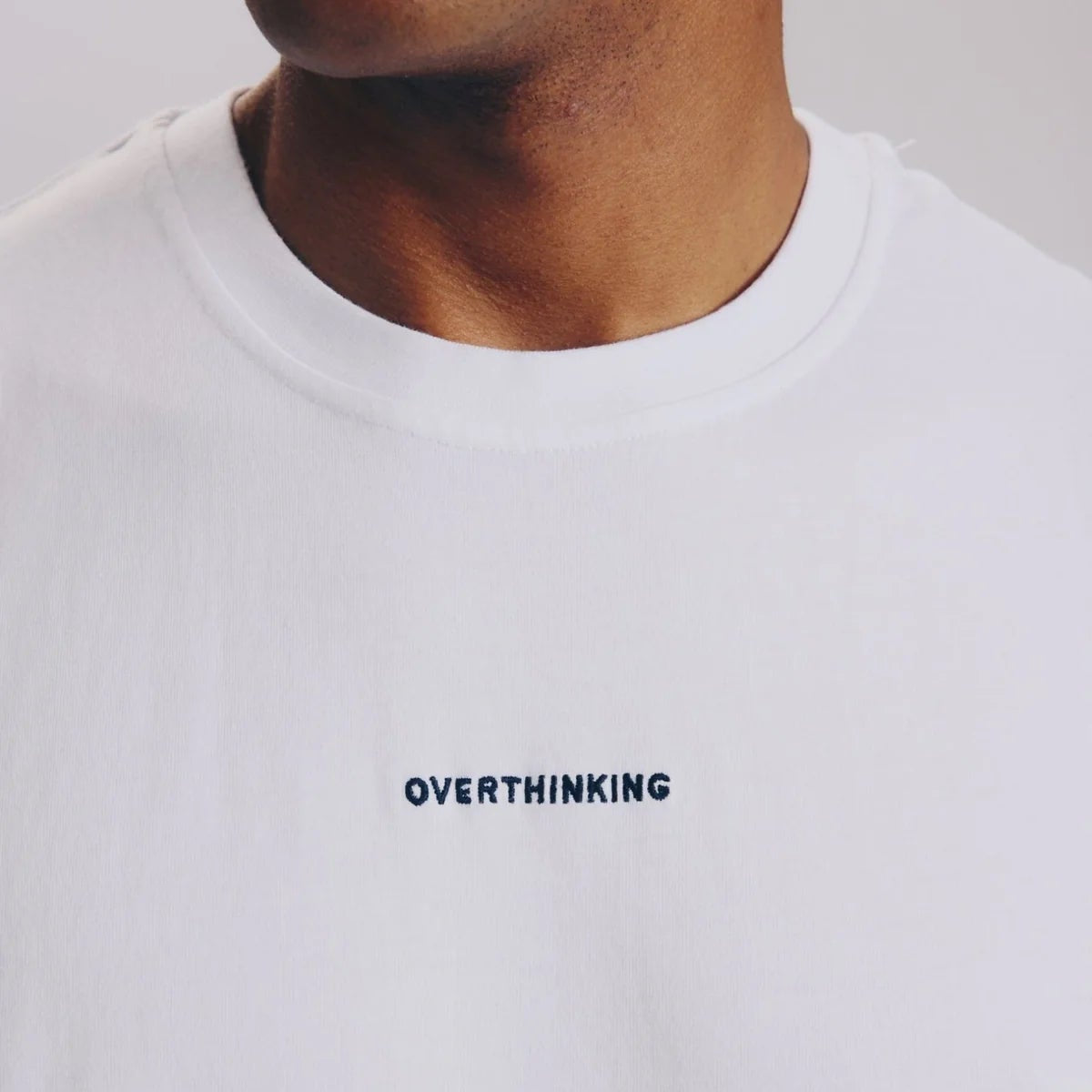 Eine Person trägt ein schlichtes weißes T-Shirt mit dem Wort OVERTHINKING, das in kleinen schwarzen Großbuchstaben auf die Brust gestickt ist. Das Gesicht der Person ist teilweise außerhalb des Rahmens.