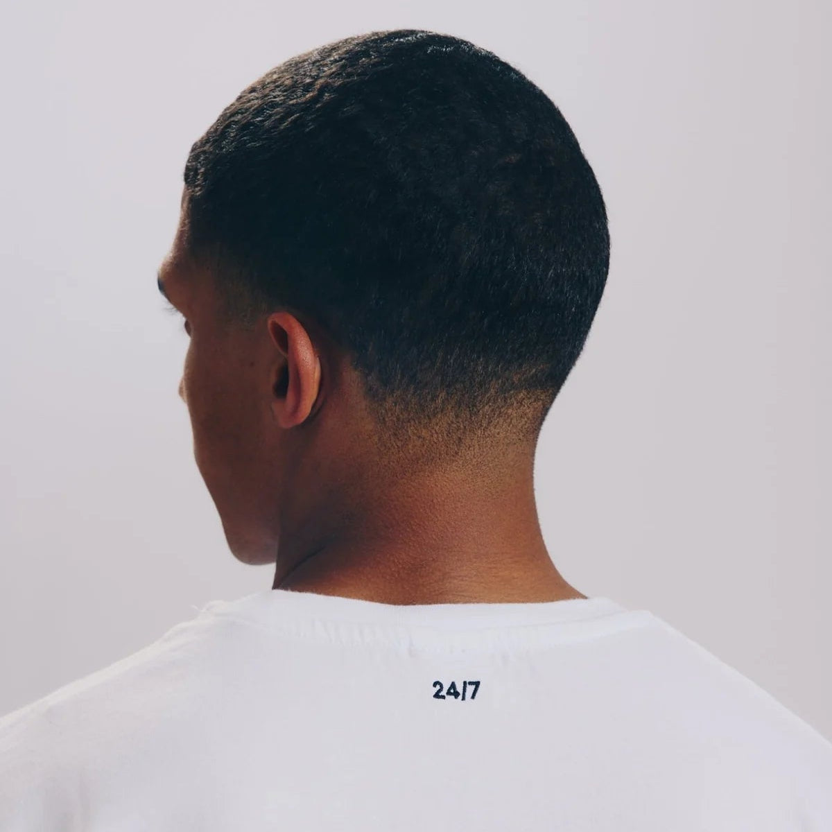 Eine Person mit kurzen, dunklen Haaren wird von hinten gezeigt. Sie trägt ein weißes Hemd mit der Aufschrift 24/7 in kleiner schwarzer Schrift auf dem Rücken nahe dem Halsausschnitt. Der Hintergrund ist einfarbig und hell.