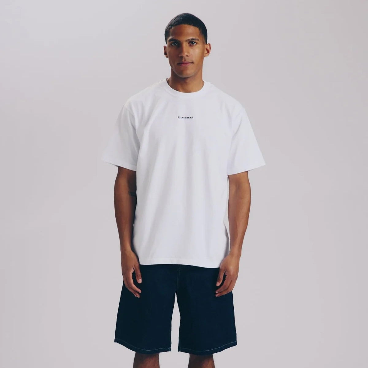 Ein Mann steht vor einem schlichten hellen Hintergrund und trägt ein lockeres weißes T-Shirt, schwarze knielange Shorts, weiße Socken und weiße Turnschuhe. Er blickt nach vorne und hat die Arme an den Seiten verschränkt.
