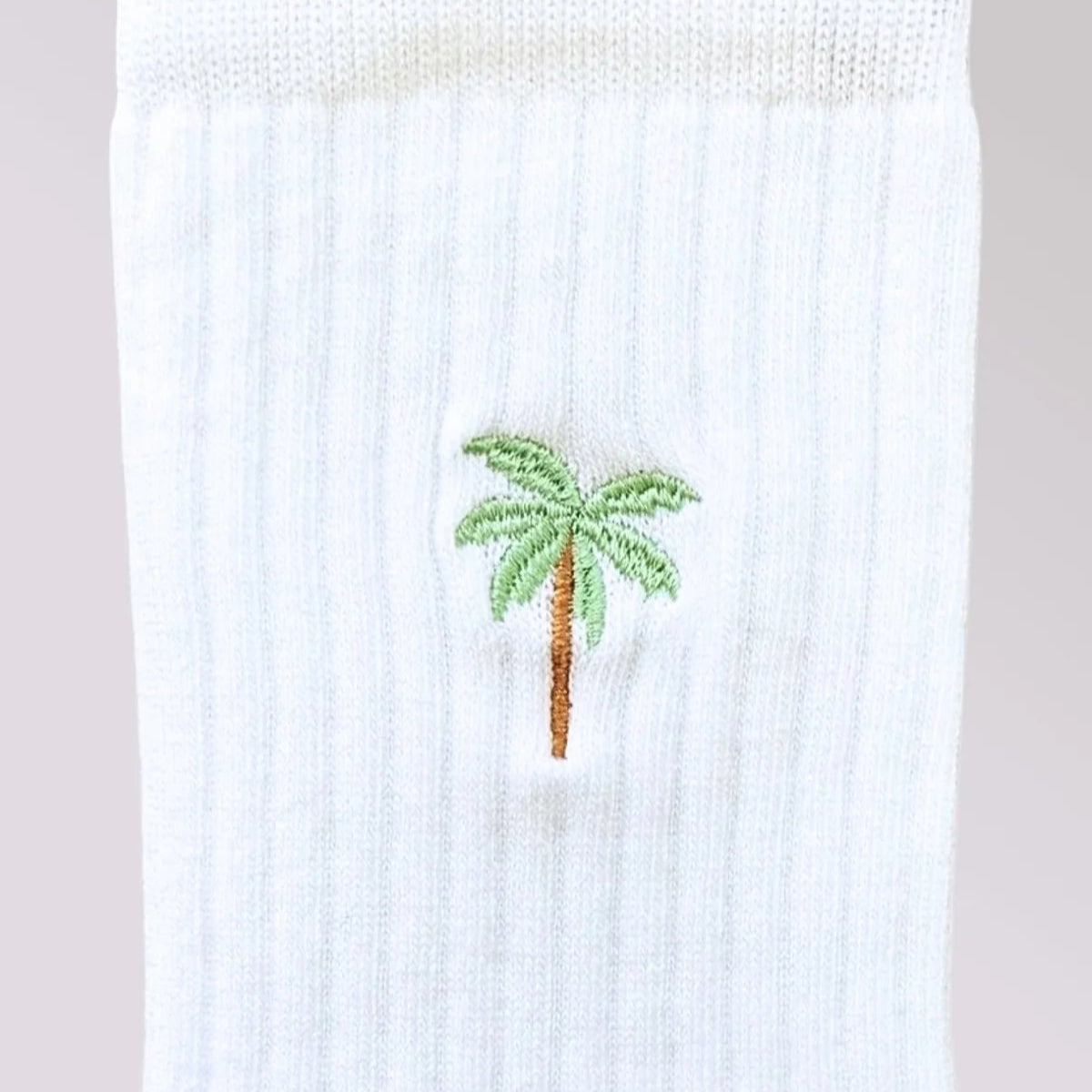 Eine Nahaufnahme einer weißen gerippten Socke mit einer kleinen gestickten grünen Palme mit braunem Stamm im Knöchelbereich.