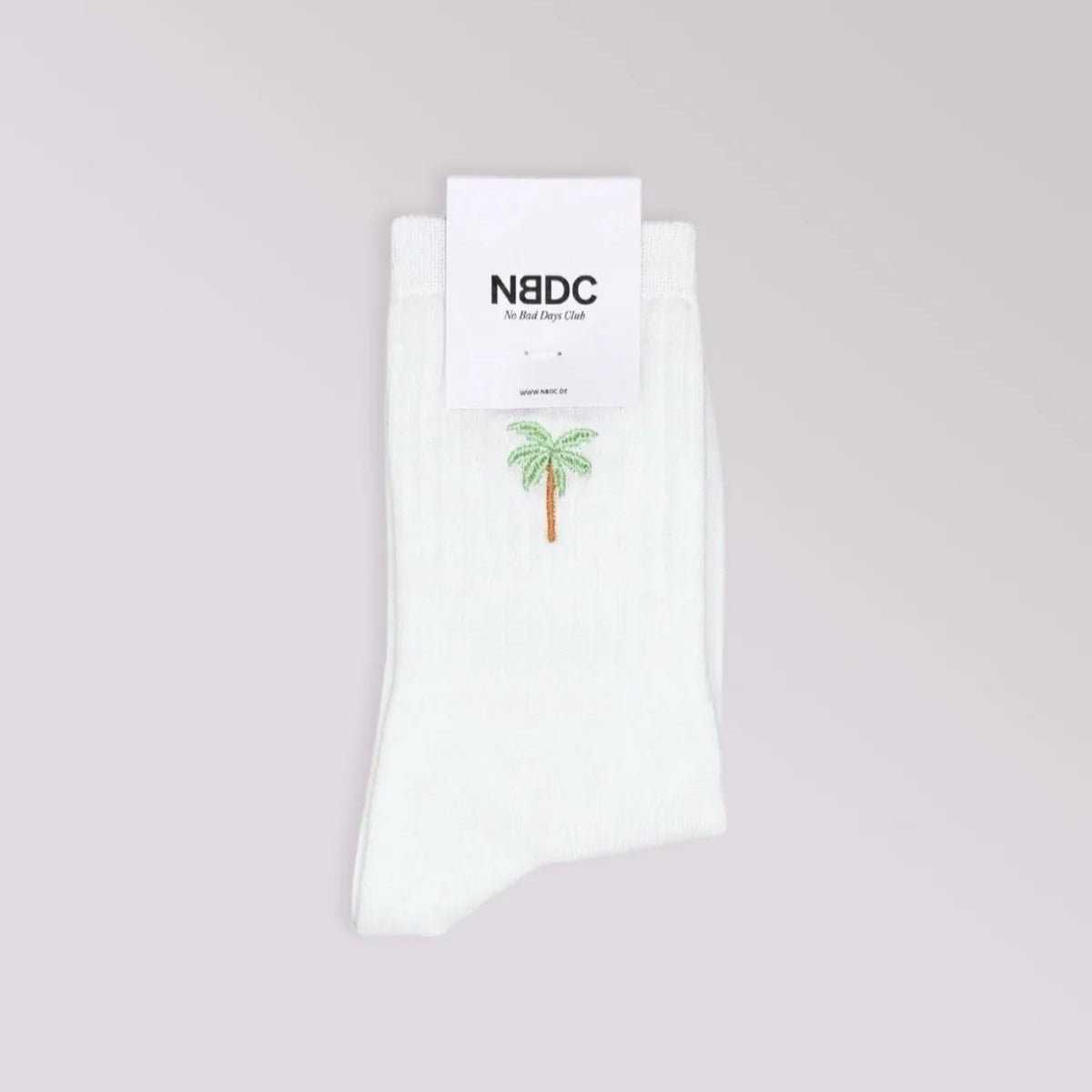 Ein Paar weiße Socken mit einer kleinen gestickten grünen Palme vor einem hellgrauen Hintergrund. Die Socken haben oben ein weißes Etikett mit dem Markennamen NBDC.