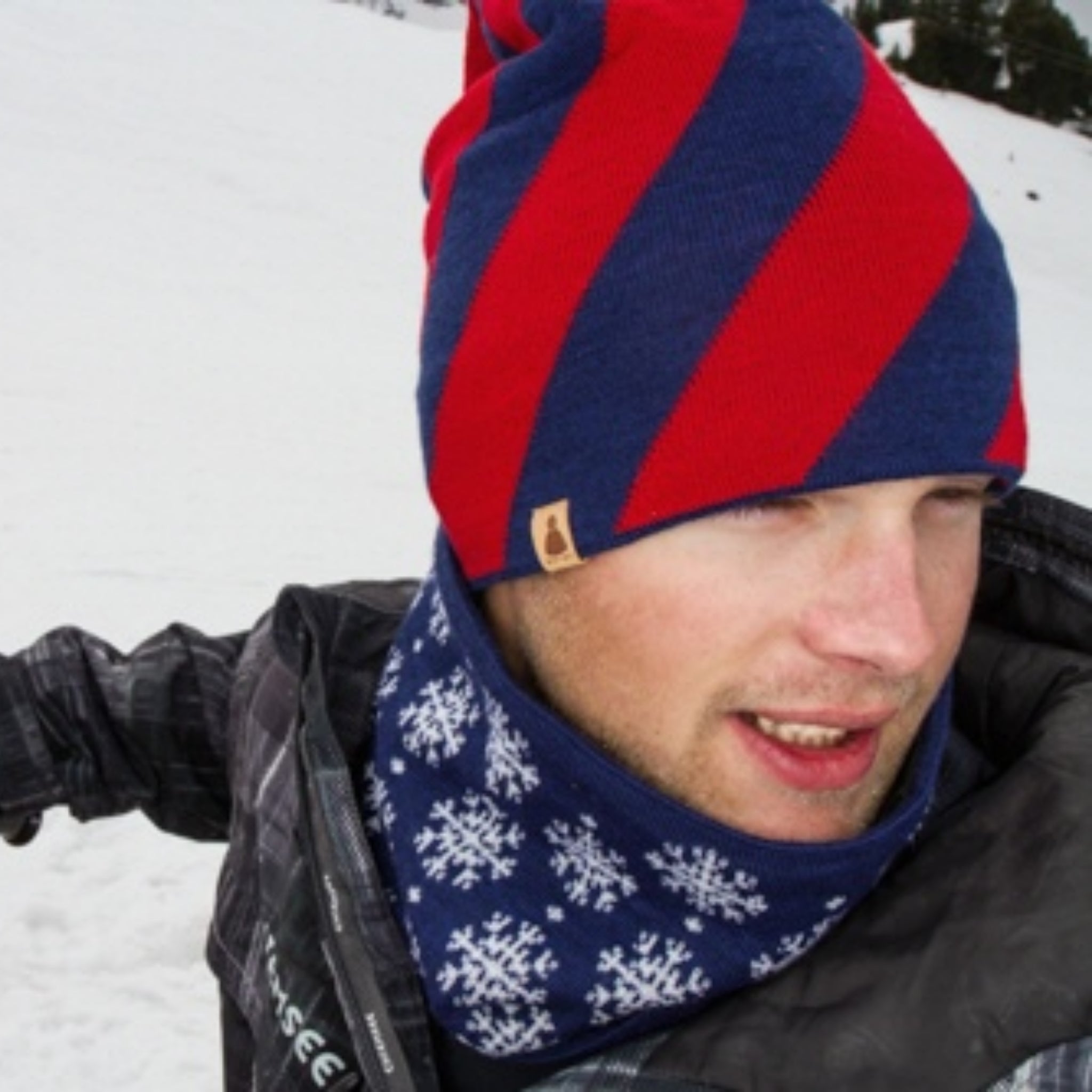 Ein Mann, der eine rot-blau gestreifte Mütze und einen blauen Schal mit weißen Schneeflocken trägt, steht in einer verschneiten Landschaft im Freien, bekleidet mit einer dunklen Winterjacke.