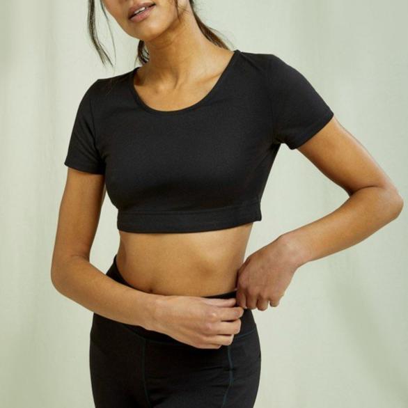 Person in schwarzem Sporttop und Hose steht vor neutralem Hintergrund und berührt den Bund der Hose.