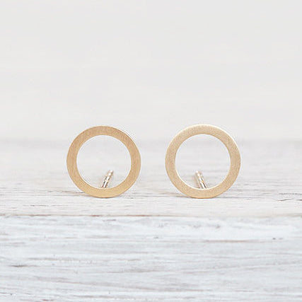 Ein Paar minimalistischer, kreisförmiger Ohrstecker aus Gold wird aufrecht auf einer hellen Holzoberfläche vor einem neutralen Hintergrund präsentiert.