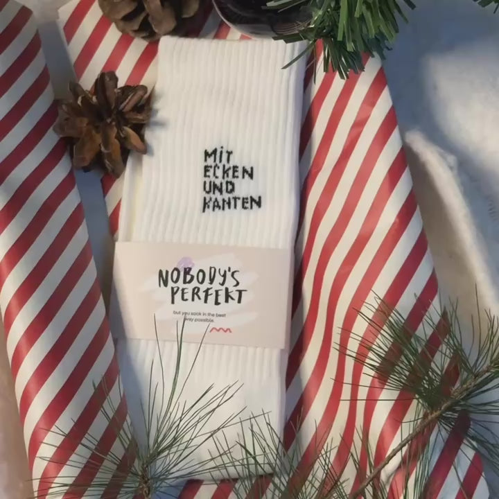 Weiße Socken mit der Aufschrift "Mit Ecken und Kanten" auf gestreiftem rot-weißem Geschenkpapier mit Kiefernadeln.