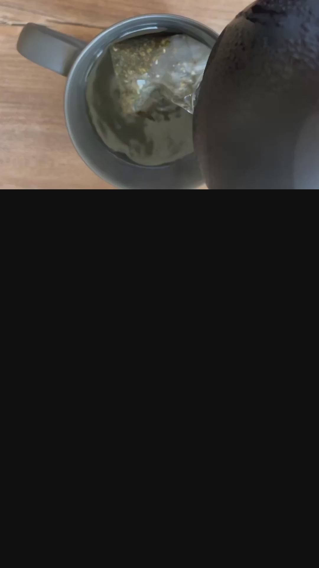 Eine schwarze Teekanne gießt heißes Wasser in eine graue Tasse mit einem Teebeutel, auf einem Holztisch.