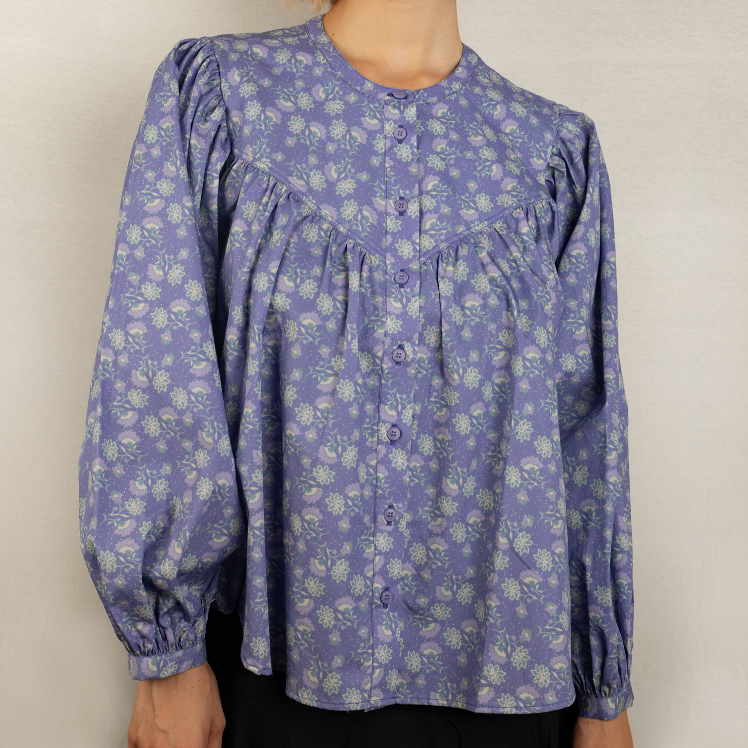 Person trägt eine lila Bluse mit Blumenmuster und langen Ärmeln vor einem neutralen Hintergrund.