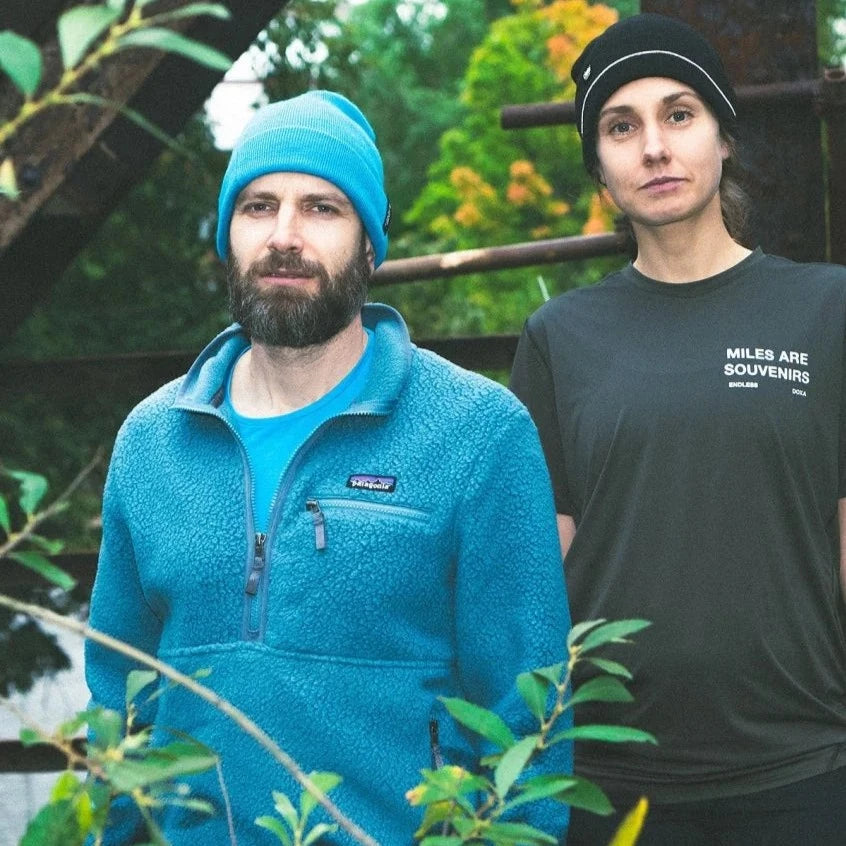 Ein Mann und eine Frau stehen draußen in Freizeitkleidung; der Mann trägt eine blaue Jacke, die Frau ein schwarzes T-Shirt.