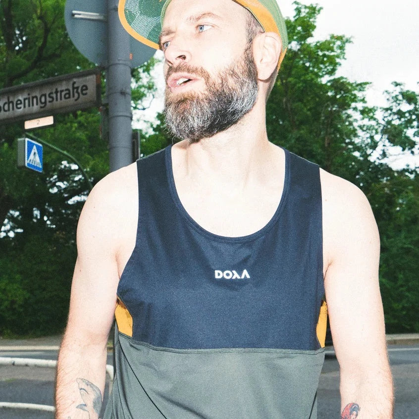 Mann mit Bart und Tanktop steht auf einer Straße neben einem Schild mit der Aufschrift "Scheringstraße", umgeben von Bäumen.