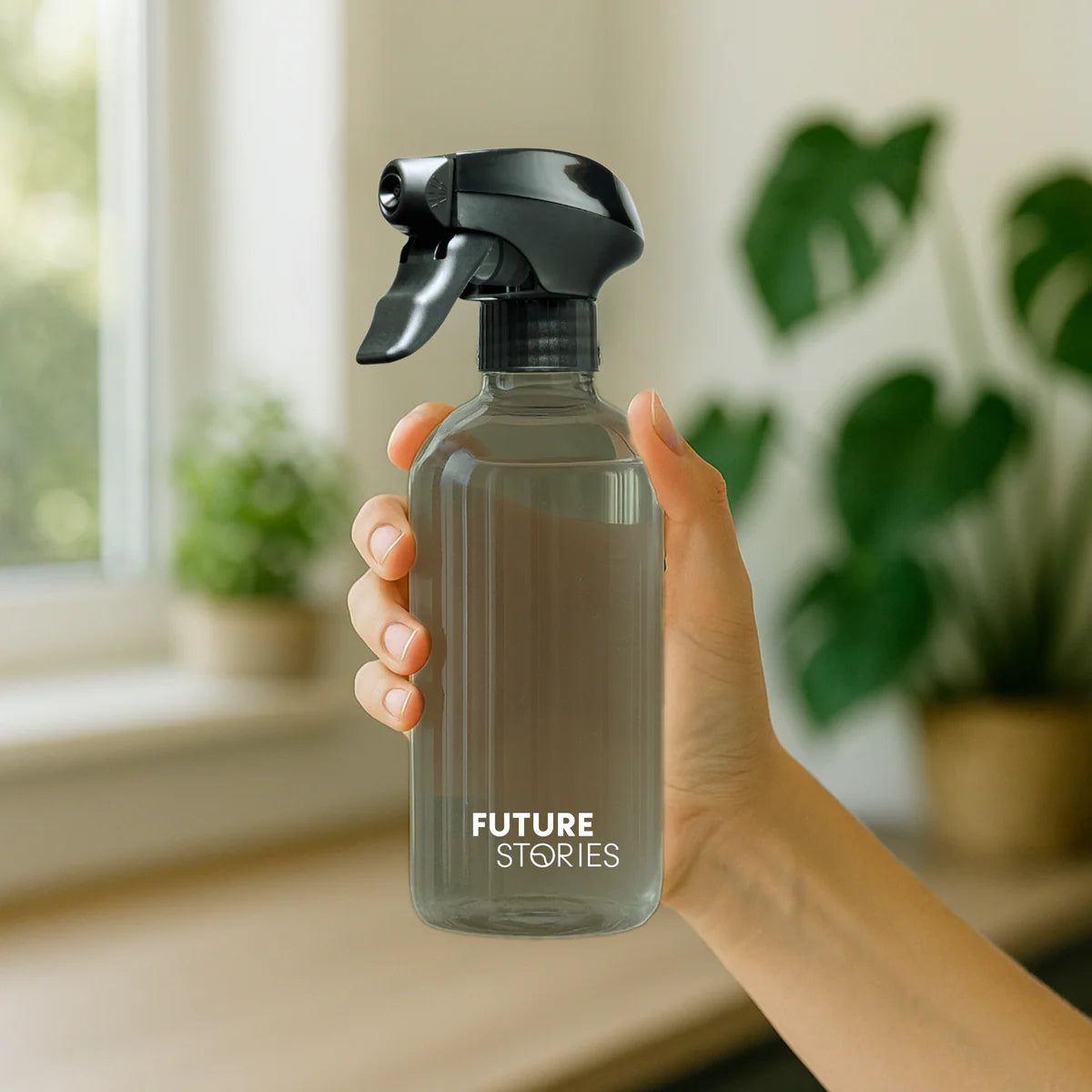 Eine Hand hält eine durchsichtige Sprühflasche mit der Aufschrift FUTURE STORIES in einem hellen Raum mit unscharfen grünen Pflanzen und einem Fenster im Hintergrund.