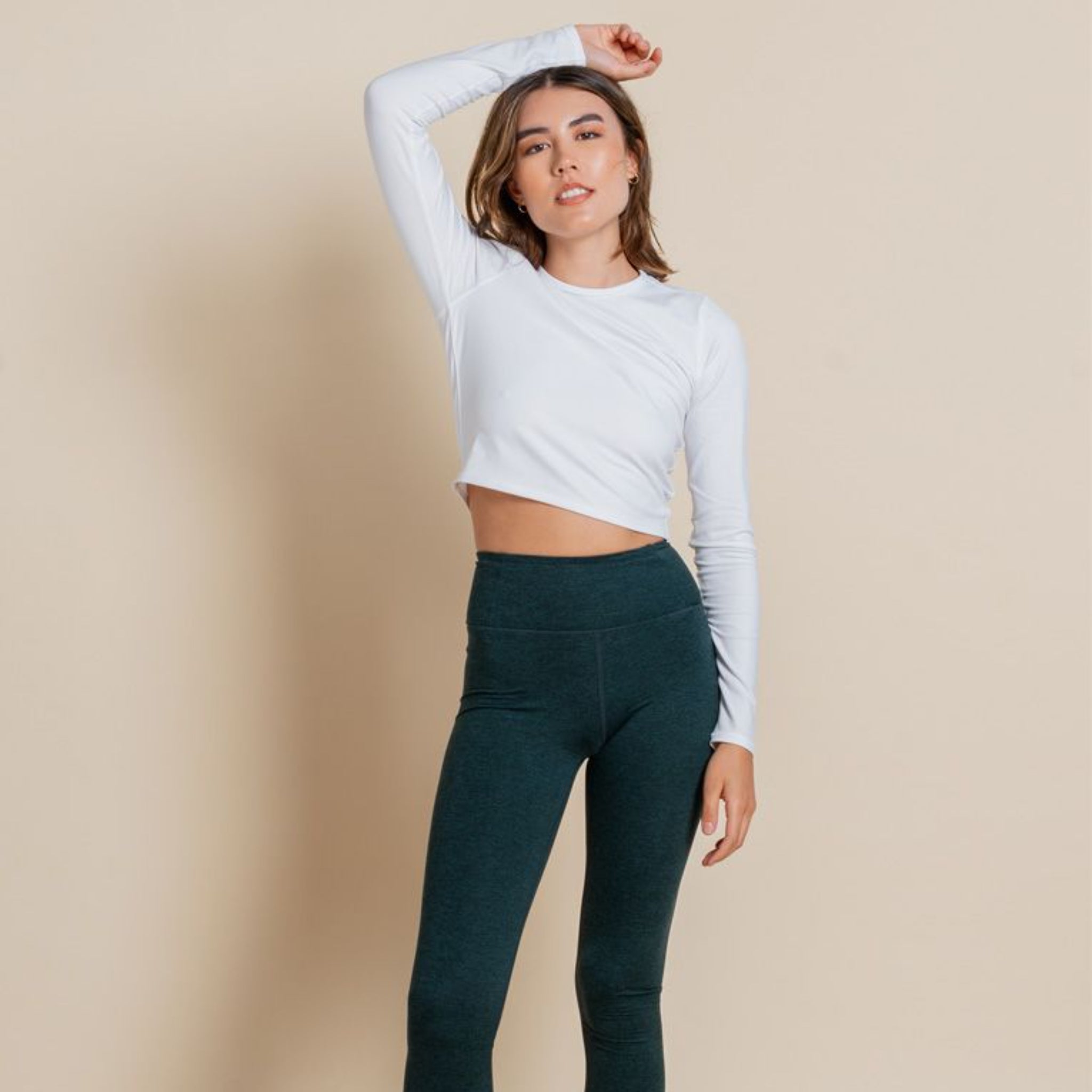 Eine Frau posiert vor einem beigen Hintergrund. Sie trägt ein weißes, langärmeliges, kurzes Oberteil und hochgezogene dunkelgrüne Leggings. Ein Arm ist am Ellbogen angewinkelt, die Hand ruht auf ihrem Kopf.