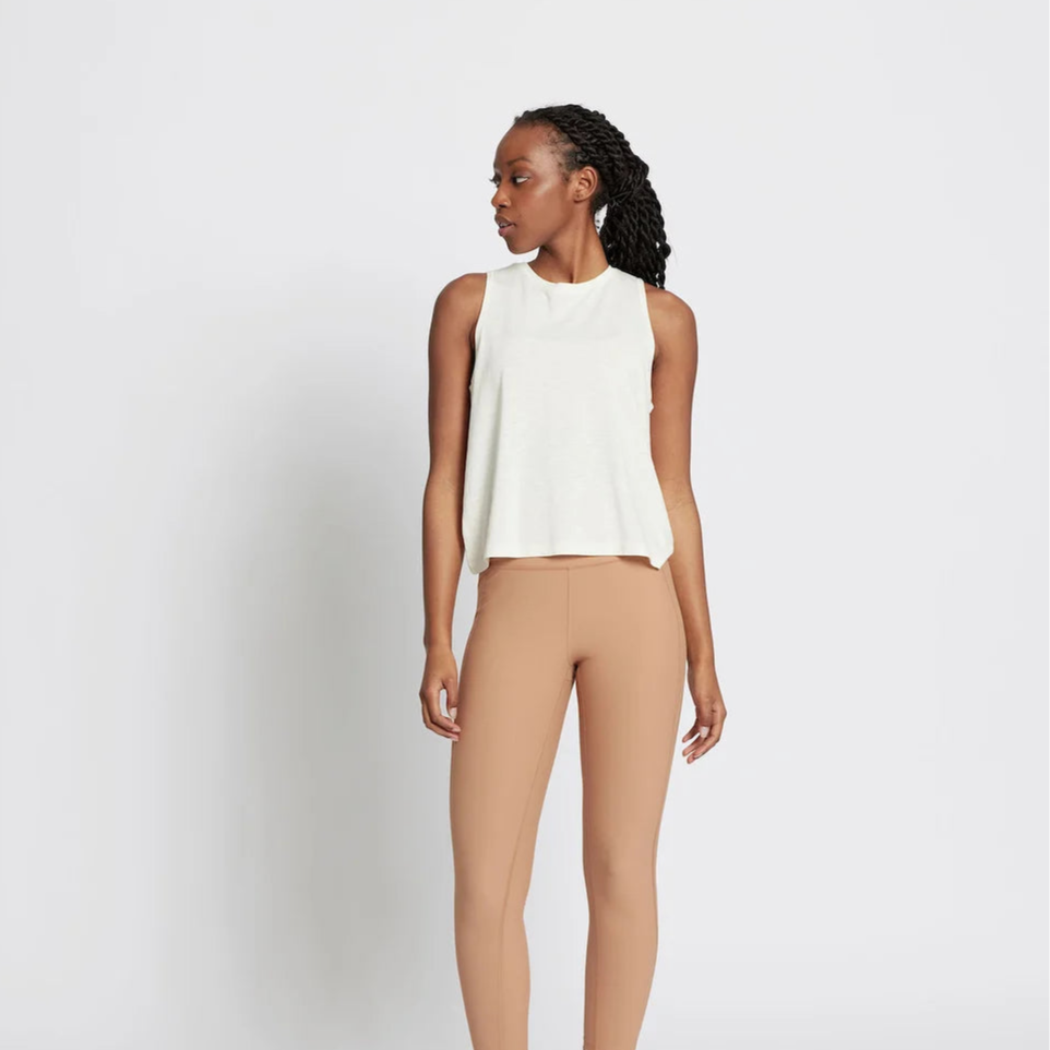 Eine Frau steht vor einem schlichten weißen Hintergrund und trägt ein ärmelloses weißes Oberteil, hellbraune Leggings mit hoher Taille und beigefarbene Turnschuhe. Sie blickt nach links und hat die Arme entspannt an der Seite.