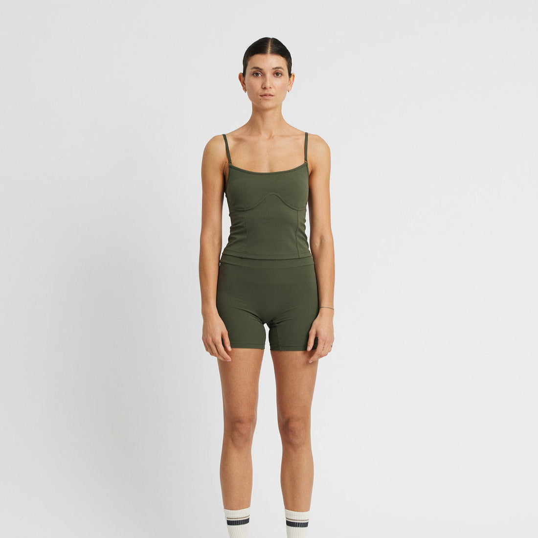 Eine Frau steht mit dem Gesicht nach vorne vor einem schlichten weißen Hintergrund. Sie trägt ein ärmelloses olivgrünes Tank-Top, dazu passende kurze Hosen, graue Socken mit schwarzen Streifen und graue Turnschuhe.