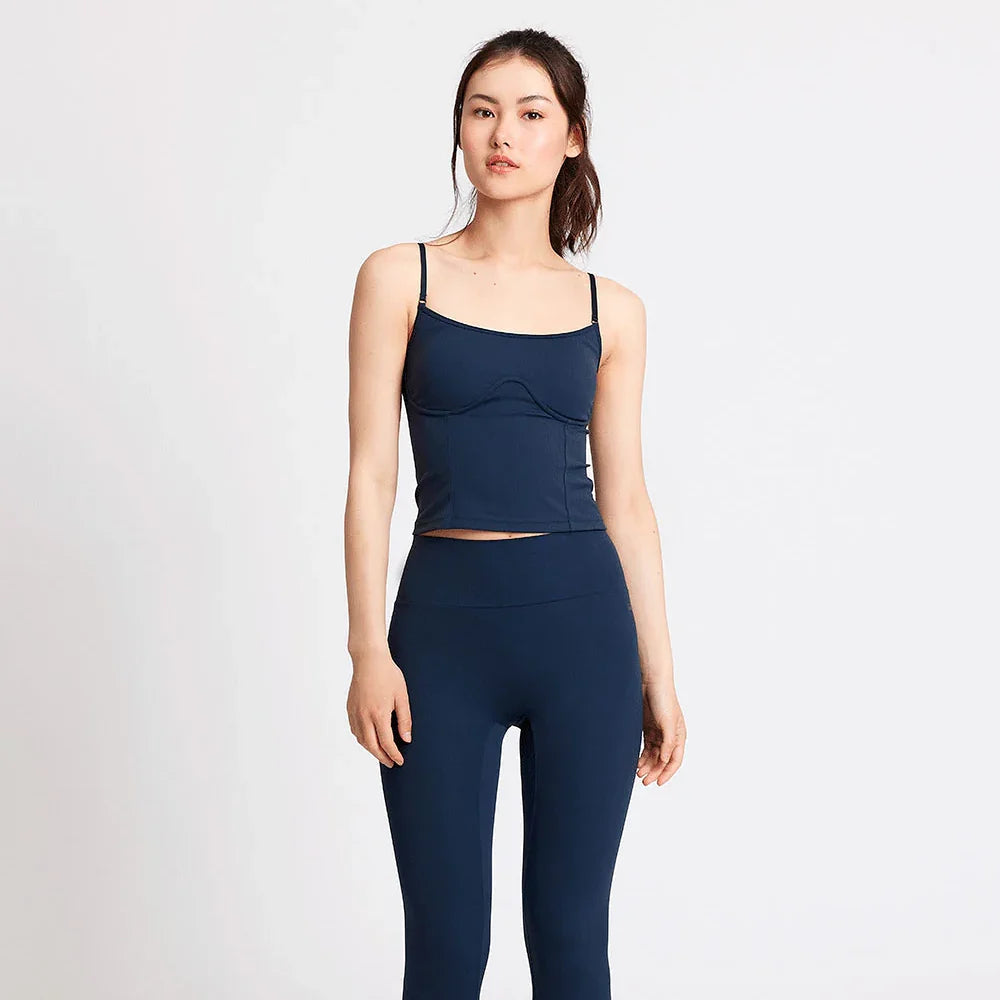 Eine Frau steht vor einem schlichten weißen Hintergrund und trägt ein marineblaues, eng anliegendes Tank-Top und passende Leggings mit hoher Taille sowie weiße Turnschuhe.