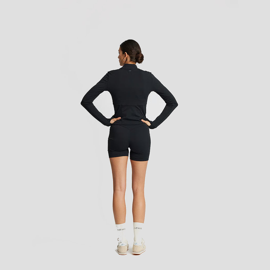 Eine Frau steht mit den Händen in den Hüften, den Blick abgewandt. Sie trägt ein schwarzes langärmeliges Sportoberteil, schwarze Shorts, weiße Socken und weiße Turnschuhe vor einem schlichten hellgrauen Hintergrund.