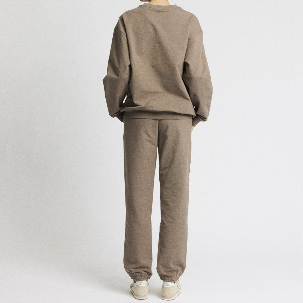 Eine Person mit dunklen Haaren in einem tief sitzenden Dutt steht mit dem Gesicht zur Seite, trägt ein locker sitzendes hellbraunes Sweatshirt und eine dazu passende Hose mit hellen Turnschuhen vor einem schlichten weißen Hintergrund.