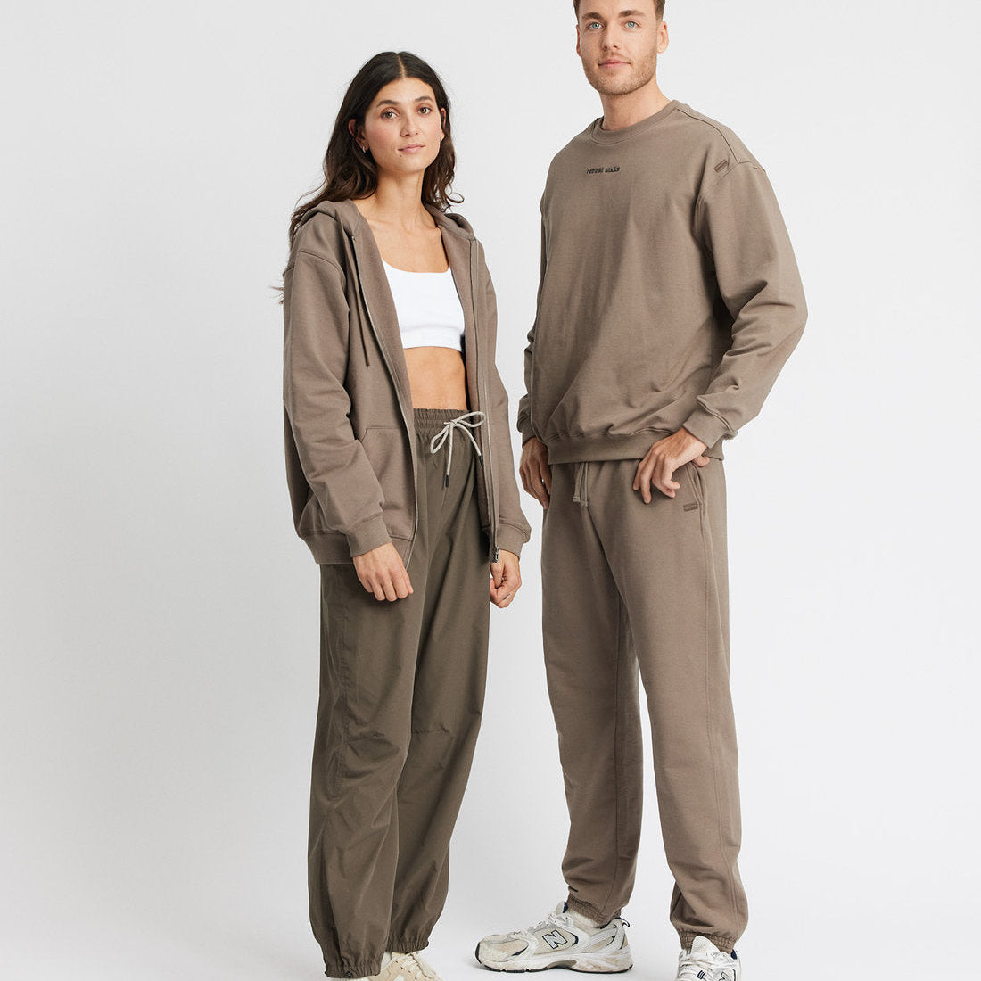 Eine Frau und ein Mann stehen nebeneinander in lässiger taupefarbener Loungewear. Die Frau trägt einen Kapuzenpulli mit Reißverschluss, ein weißes Crop-Top und Jogginghosen, der Mann ein Sweatshirt und Jogginghosen. Beide tragen Turnschuhe und schauen nach vorne.