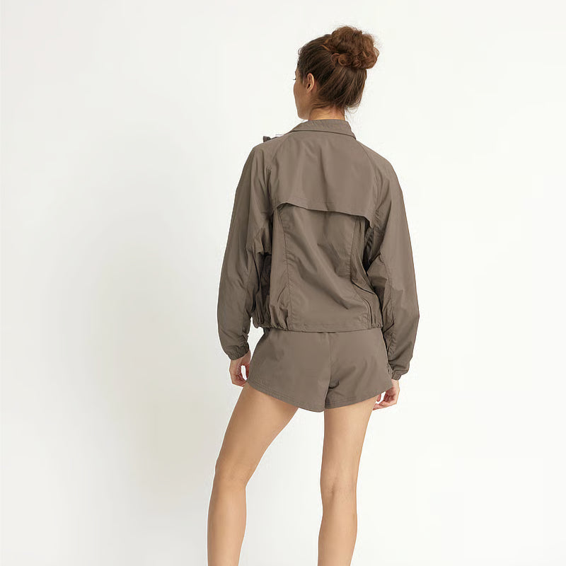 Eine Frau steht von der Kamera abgewandt und trägt eine taupefarbene Windjacke und passende Shorts. Sie hat ihr Haar zu einem Dutt gebunden und posiert vor einem schlichten weißen Hintergrund.