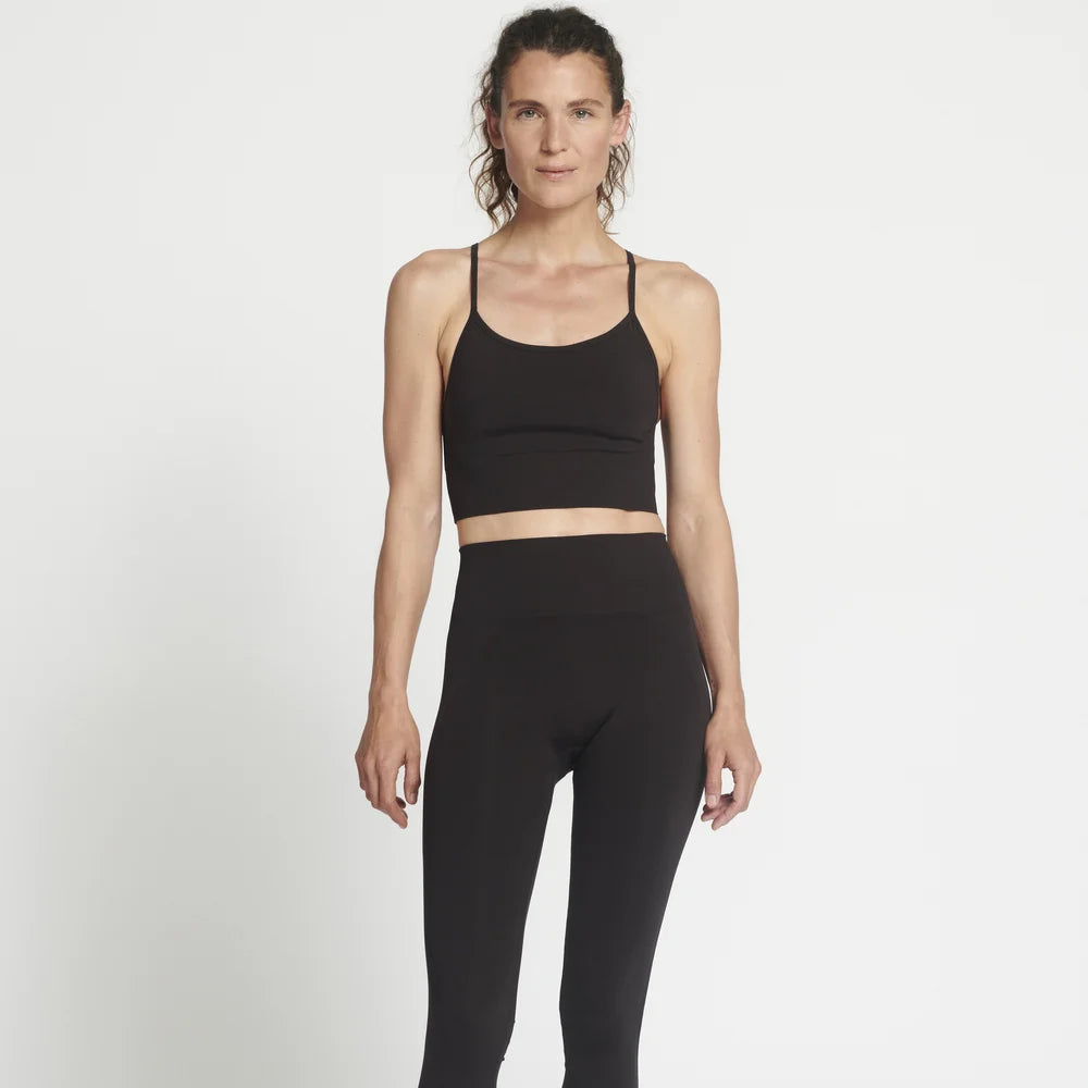 Eine Frau steht barfuß vor einem schlichten weißen Hintergrund und trägt einen schwarzen Sport-BH und schwarze Leggings mit hoher Taille. Sie hat ihr lockiges Haar zurückgebunden und einen neutralen Gesichtsausdruck.