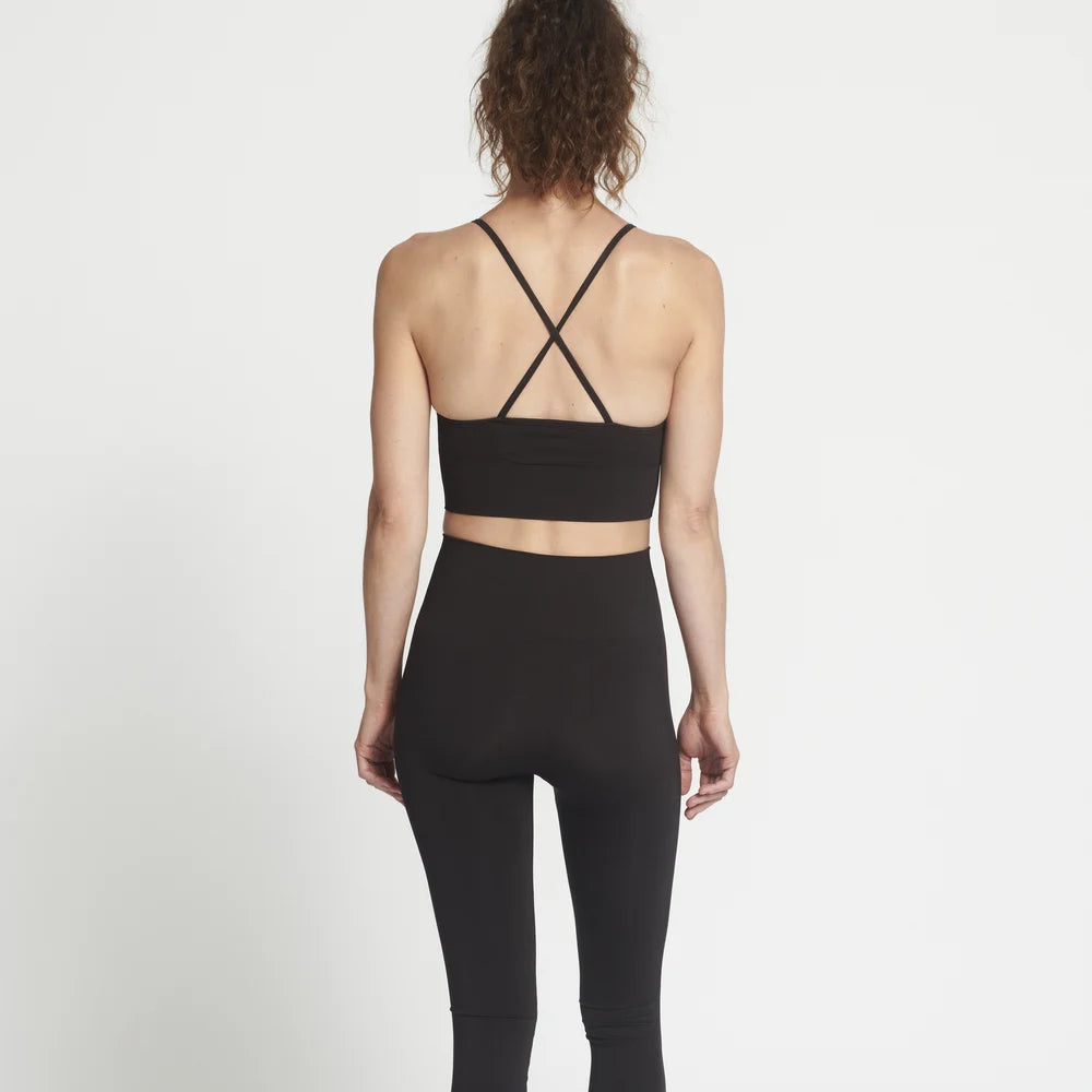 Eine Frau steht barfuß mit dem Gesicht zur Wand und trägt einen schwarzen Sport-BH mit überkreuzten Trägern und dazu passende hoch taillierte Leggings vor einem schlichten weißen Hintergrund.