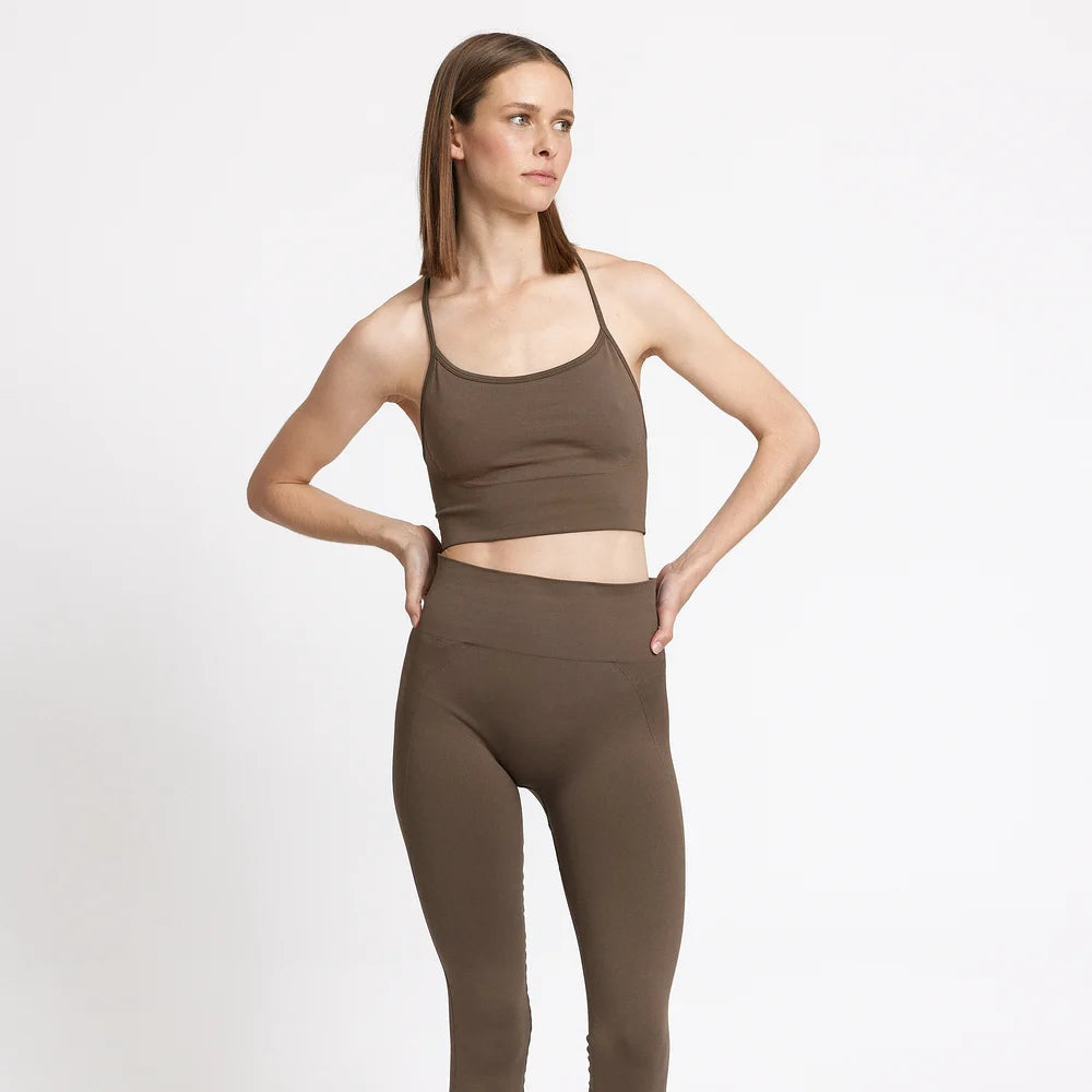 Eine Frau steht barfuß vor einem schlichten weißen Hintergrund und trägt einen braunen Sport-BH und dazu passende Leggings mit hoher Taille. Sie hat glattes braunes Haar und blickt mit einer Hand in der Hüfte zur Seite.