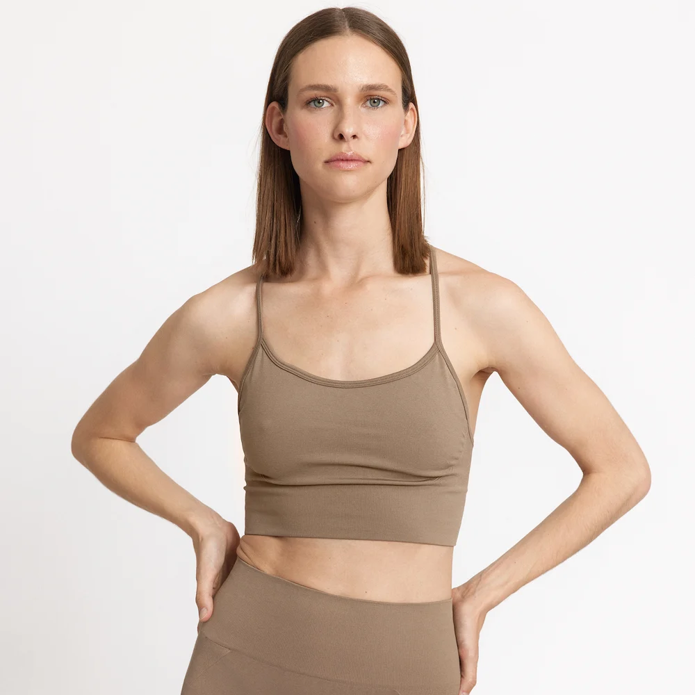 Eine Frau mit glattem braunem Haar trägt einen passenden taupefarbenen Sport-BH und hoch taillierte Leggings und steht mit einer Hand auf der Hüfte vor einem schlichten weißen Hintergrund.