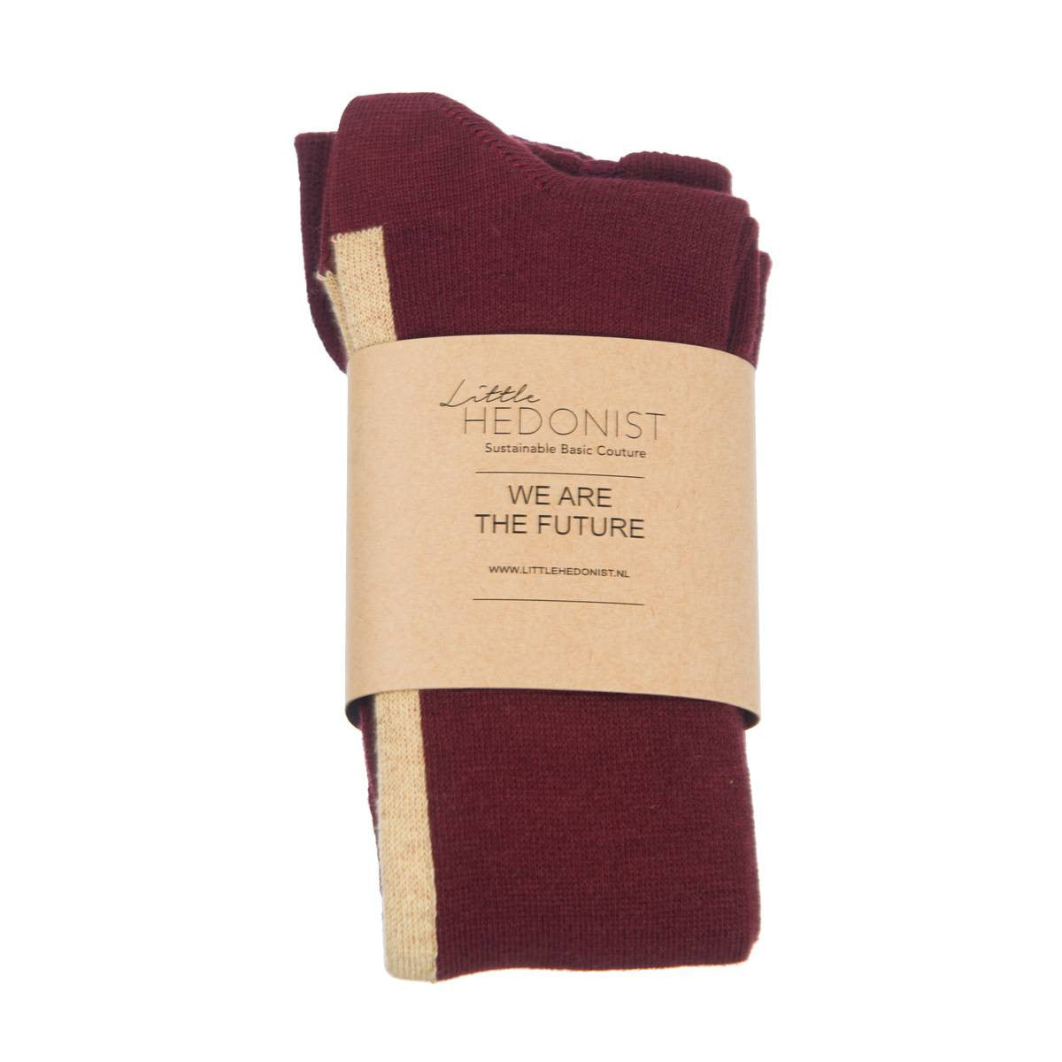 Ein Paar gefaltete kastanienbraune Socken mit einem beigen Streifen, eingewickelt in ein braunes Papierband mit der Aufschrift Little Hedonist Sustainable Basic Couture. Wir sind die Zukunft.