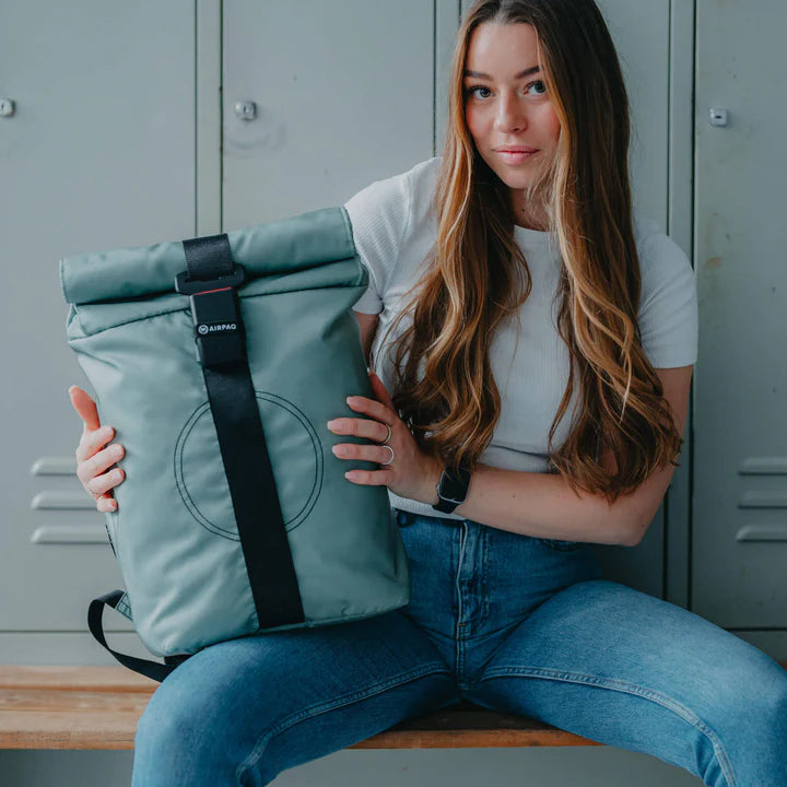 Eine Frau mit langen braunen Haaren, die ein weißes Oberteil und blaue Jeans trägt, sitzt auf einer Bank vor grauen Spinden und hält einen hellgrünen Rolltop-Rucksack auf dem Schoß.