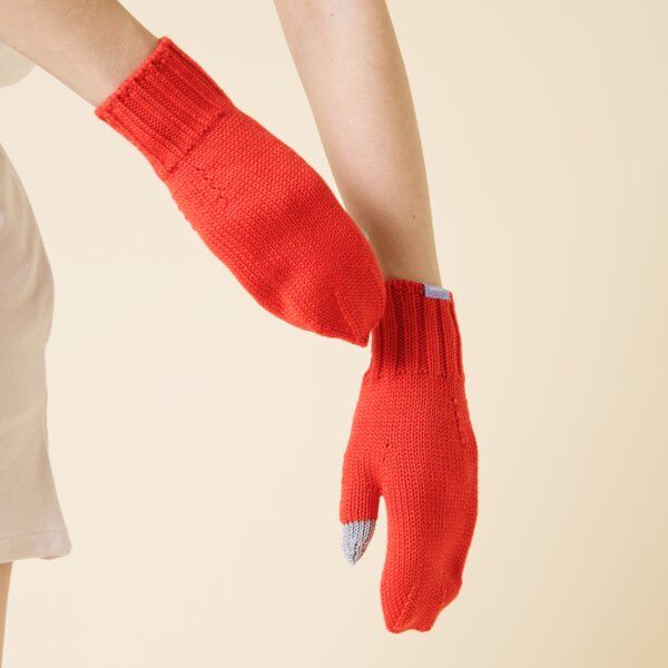 Eine Person, die leuchtend rote Strickfäustlinge trägt, wobei ein Fäustling eine graue Daumenspitze aufweist, streckt ihren Arm vor einem einfarbig beigen Hintergrund aus. Nur ein Teil des hellen Outfits der Person ist sichtbar.
