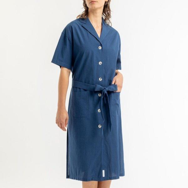 Person trägt ein blaues Hemdkleid mit Knöpfen und einem Gürtel, stehend vor einem weißen Hintergrund.