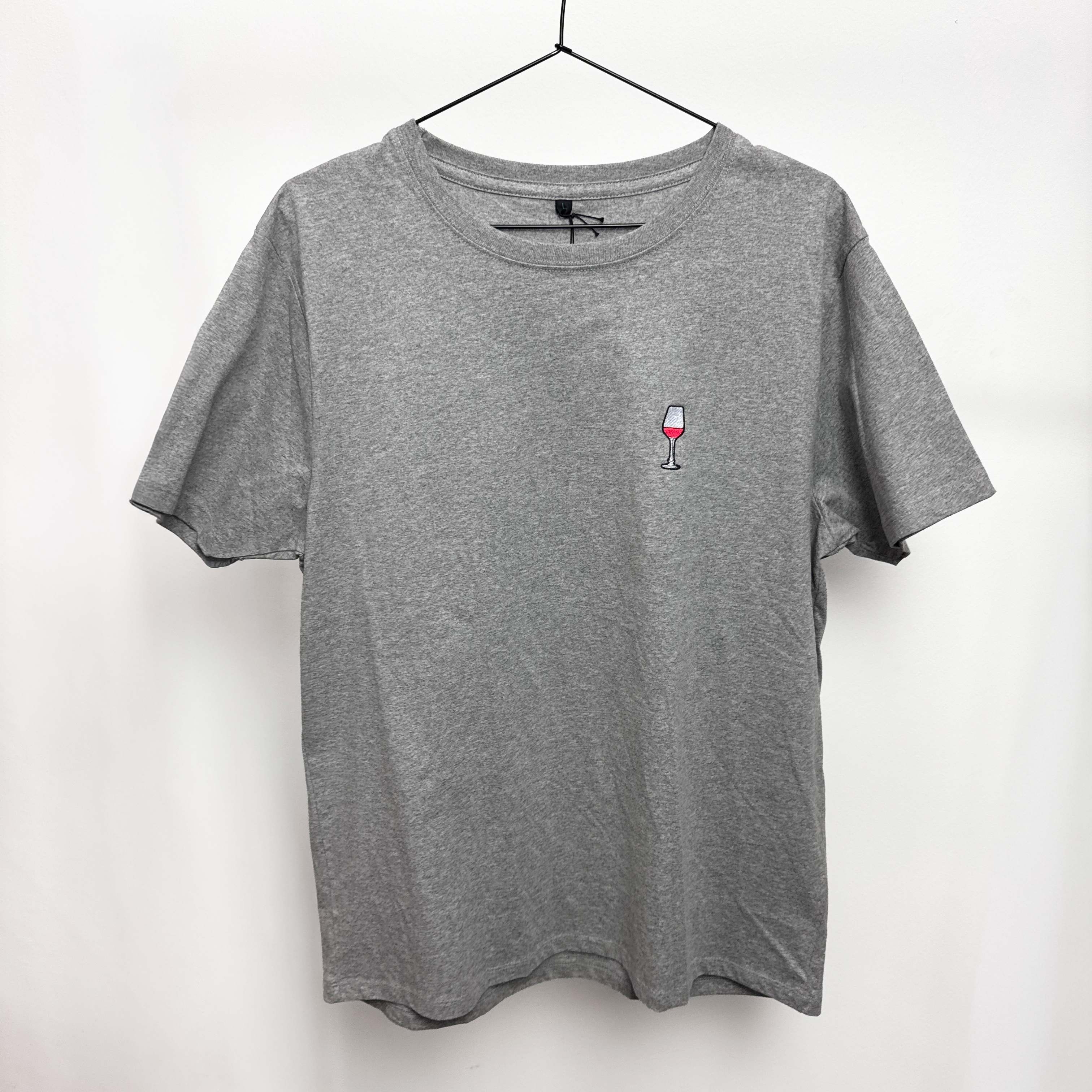 Ein schlichtes graues T-Shirt auf einem schwarzen Bügel, mit einem kleinen gestickten Motiv eines mit Rotwein gefüllten Weinglases auf der linken Brustseite.