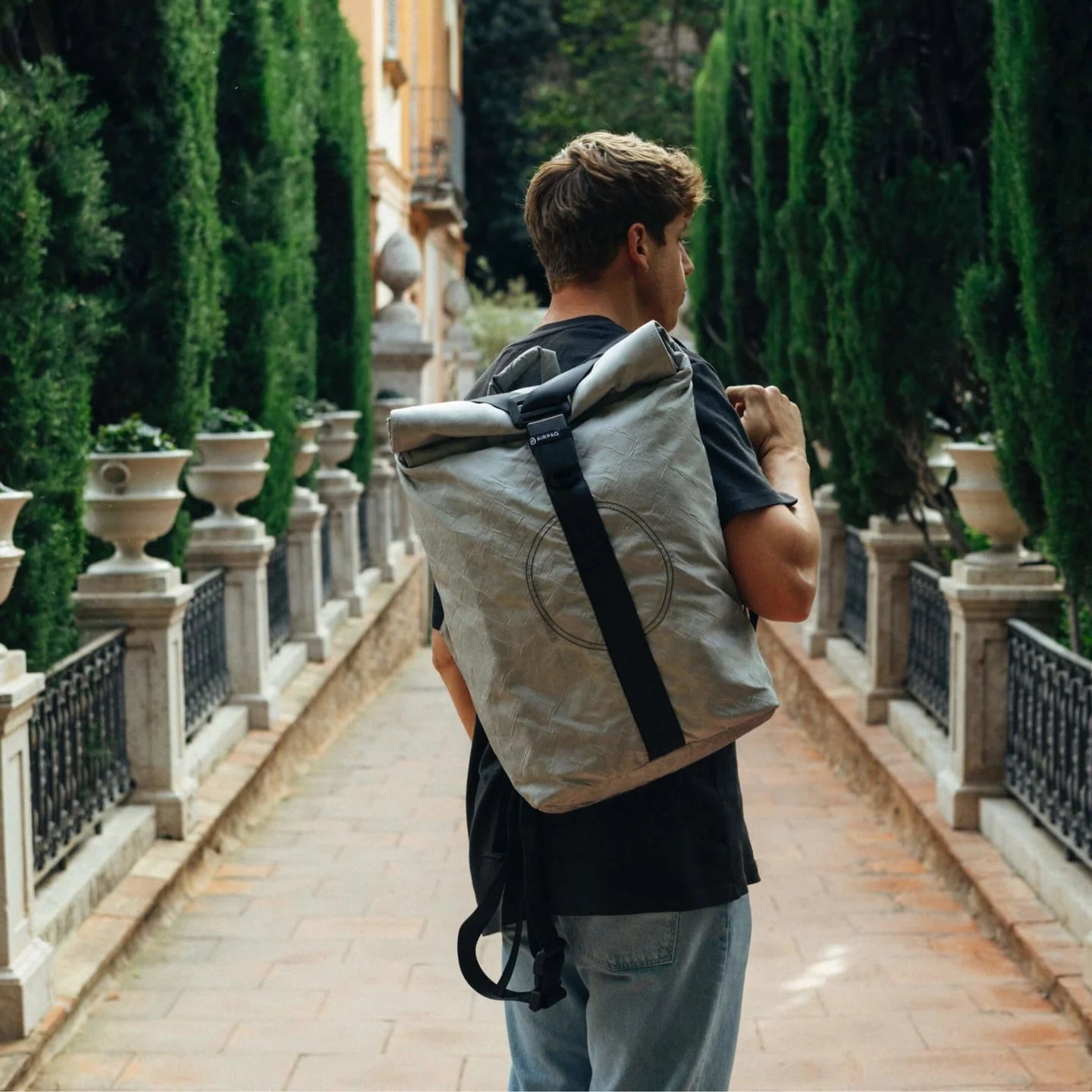 Ein Mann mit grauem Rucksack geht auf einem gepflasterten Weg mit hohen Hecken und Topfpflanzen.
