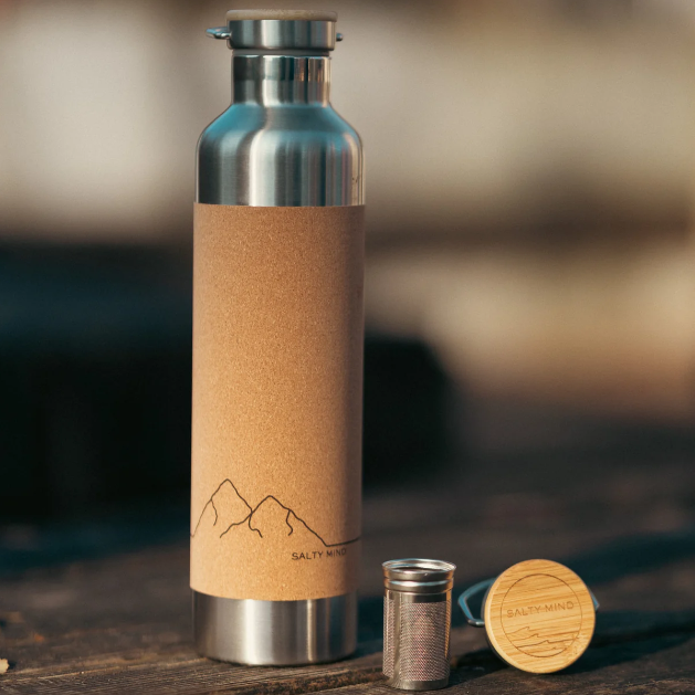 Eine Wasserflasche aus Edelstahl mit Korkmanschette und minimalistischem Bergdesign, die zusammen mit ihrem Bambusdeckel und einem Metallsieb auf einer Holzfläche steht.