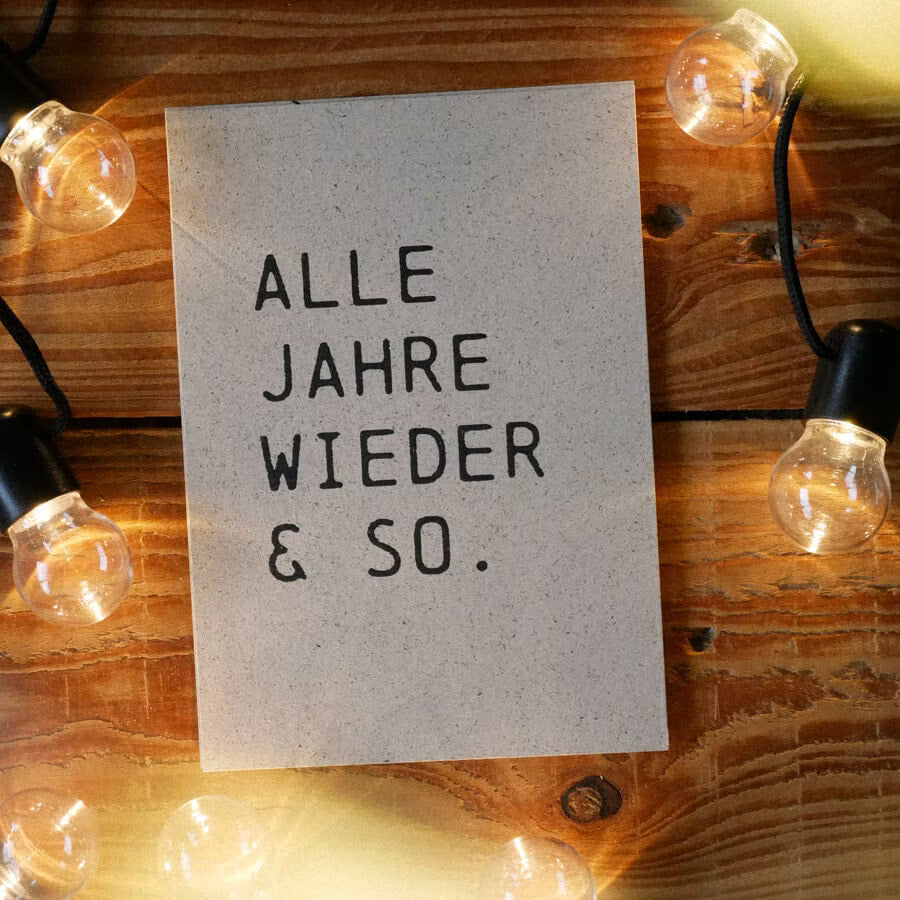 Karte mit dem Text "ALLE JAHRE WIEDER & SO." auf Holzuntergrund, umgeben von Glühbirnen.