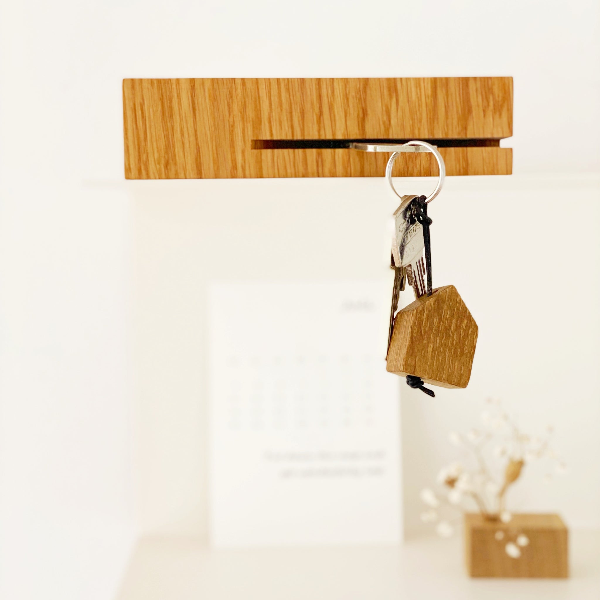 Ein Schlüsselbund mit einem kleinen Holzhaus-Schlüsselanhänger hängt an einem hölzernen Halter an einer weißen Wand, mit einem verschwommenen Kalender und einer dekorativen Pflanze im Hintergrund.